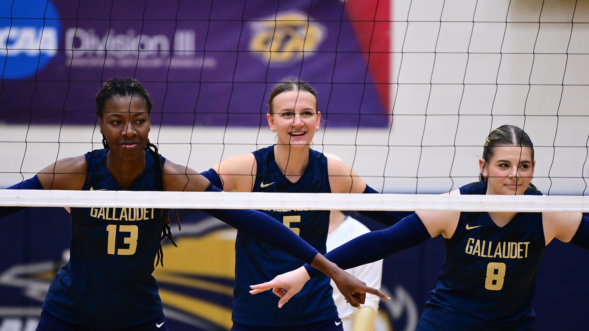 Gallaudet women’s volleyball players Kayla Debrow (#13), Jada Zarembka (#5) and Aliyah Medearis (#8), from left to right, stand at the net in their navy Bison uniforms with arms extended and hands pointing as they communicate and prepare for the next play during a match in the Field House.