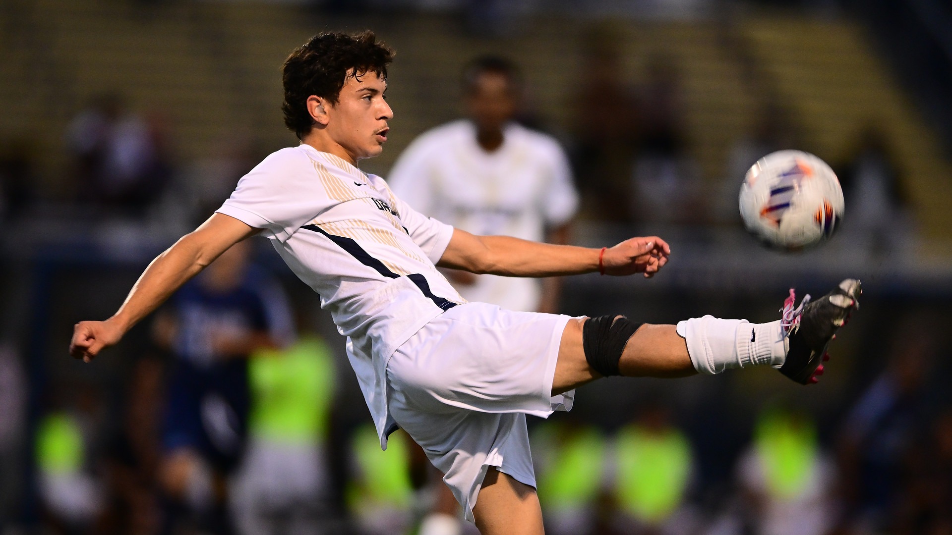 Gallaudet men’s soccer player Daniel Maye (#8) extends his right leg high into the air to kick a soccer ball during a night match at Hotchkiss Field. Maye wears a white-and-gold Bison uniform as teammates and spectators appear blurred in the background.