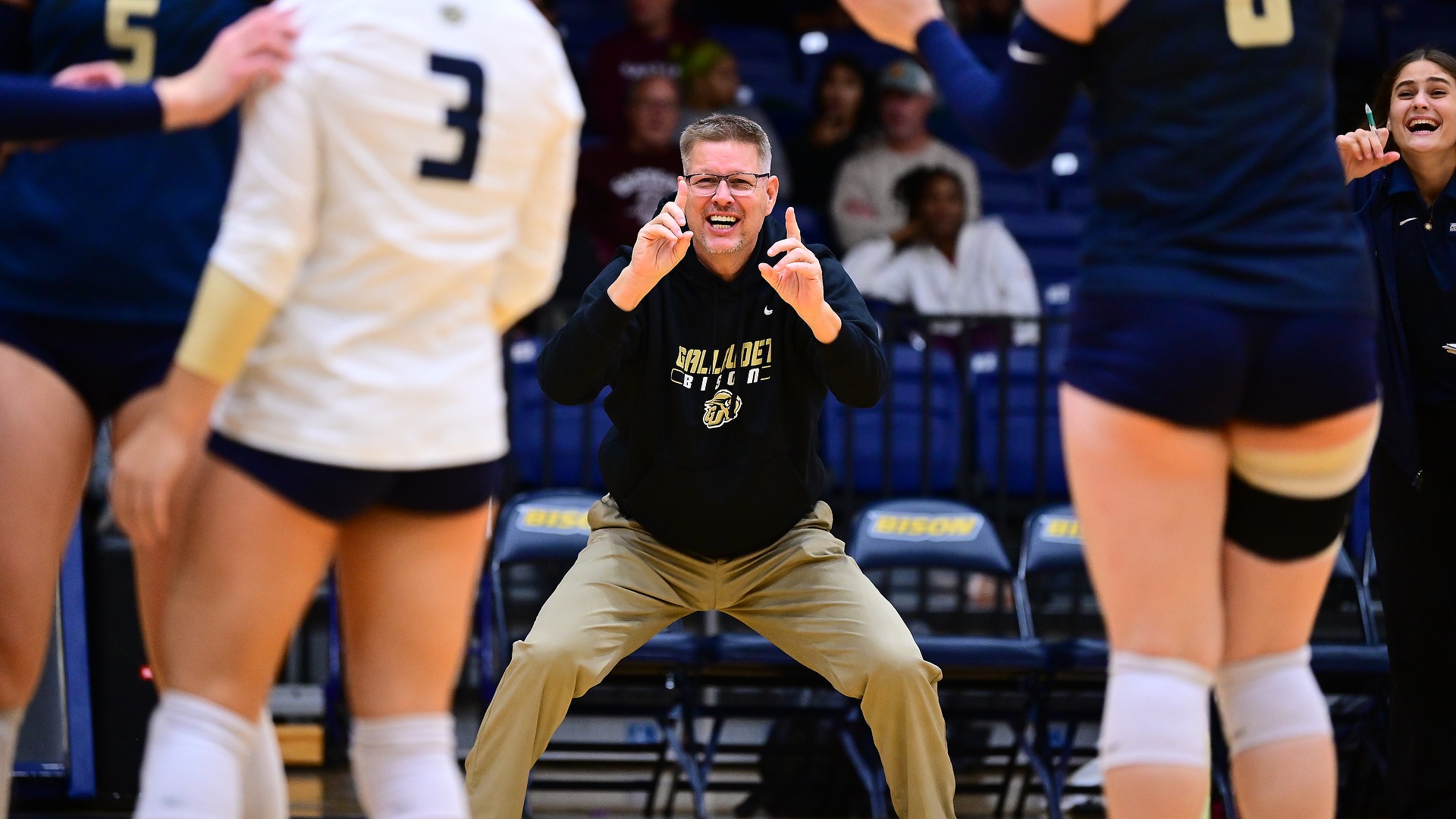 Gallaudet University women's volleyball head coach Lynn Ray Boren crouches along the sideline in a black Bison hoodie and khaki pants, using American Sign Language (ASL) to communicate instructions to his players during a match in the Field House. Bison players in navy-and-white uniforms frame the foreground, while an assistant coach cheers in the background.