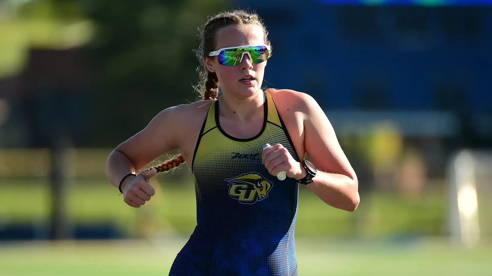 Gallaudet University women's triathlon student-athlete Alecia Miller runs during the Bison's home AquaBike event on Sept. 6, 2026. Miller wears a navy-and-gold Gallaudet triathlon suit featuring the Bison logo, reflective green sunglasses and a sports watch, with her braided hair trailing behind her as she runs in the morning sunlight.