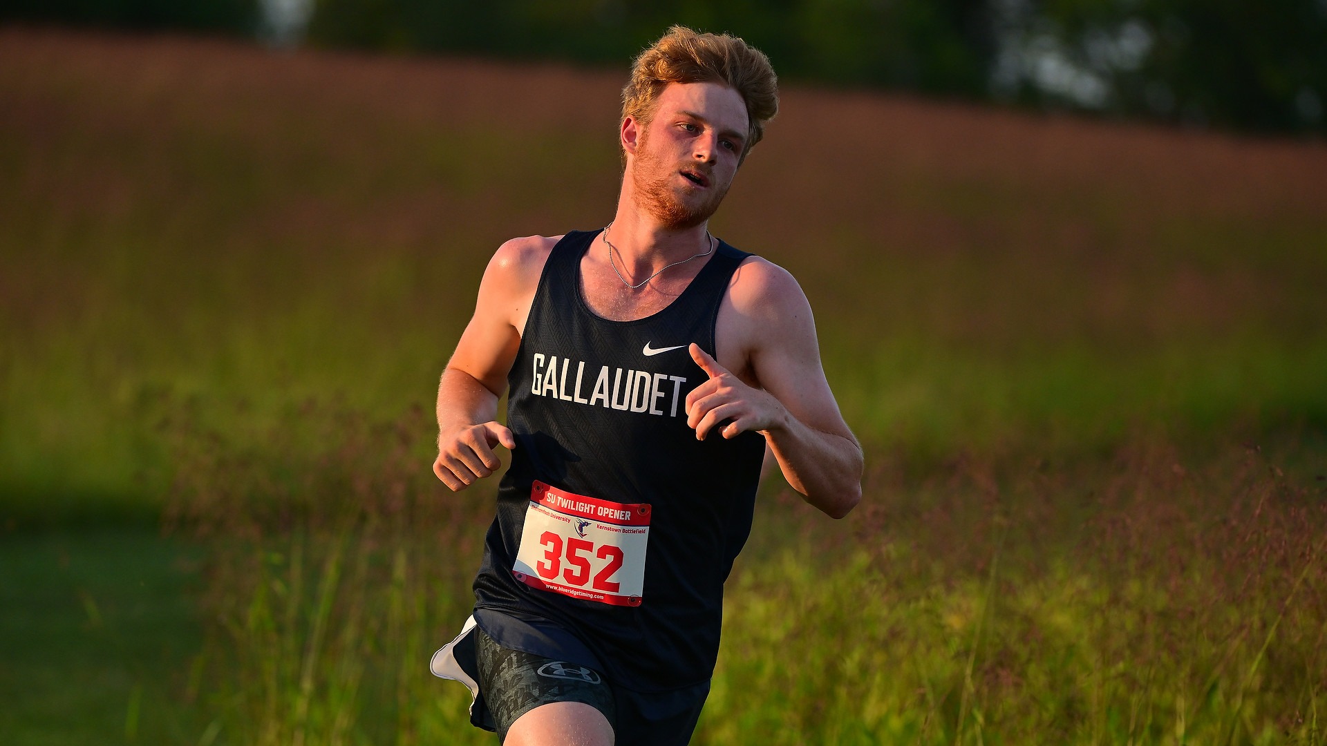Gallaudet University men's cross country runner Troy Holcomb runs along a grassy course during the Shenandoah Twilight Opener on Sept. 11, 2026. Holcomb wears a navy Gallaudet racing singlet, navy shorts and race bib No. 352 as the warm evening sunlight illuminates his face and hair, with a grassy hillside blurred in the background.