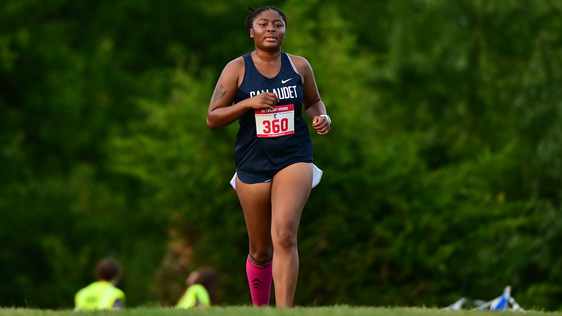 Gallaudet University women's cross country runner Jala Furdge runs alone along a grassy course during the Shenandoah Twilight Opener on Sept. 11, 2026. Furdge wears a navy Gallaudet uniform, race bib No. 360, a bright pink compression sleeve on her right calf and a smartwatch, with lush green trees and race officials blurred in the background.