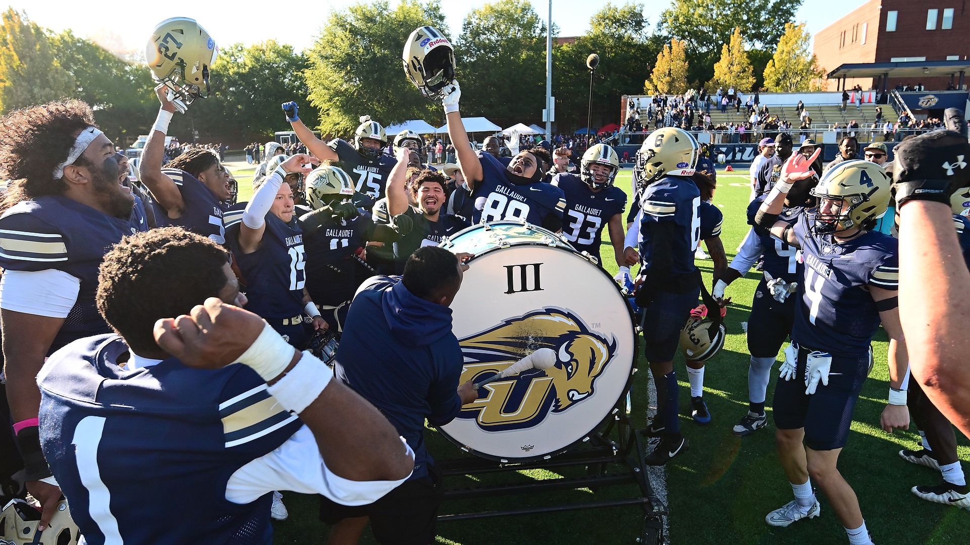Gallaudet University football players gather around the large Bison drum at midfield for their traditional postgame celebration following a 45-34 Homecoming victory over the Coppin State University club football team on Oct. 17 at Hotchkiss Field. Players in navy-and-gold uniforms cheer, raise their helmets and pump their fists as a team member pounds the drum, celebrating the win on a sunny fall afternoon in Washington, D.C.