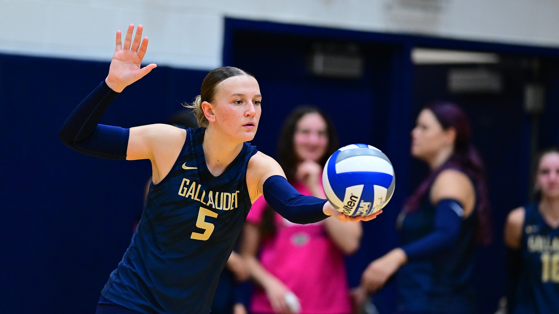 Gallaudet women's volleyball player Jada Zarembka (#5) prepares to serve during a match, holding a blue, white and gray volleyball in her left hand while raising her right hand above her head. Zarembka wears a navy-and-gold Bison uniform, with teammates and spectators blurred in the background.