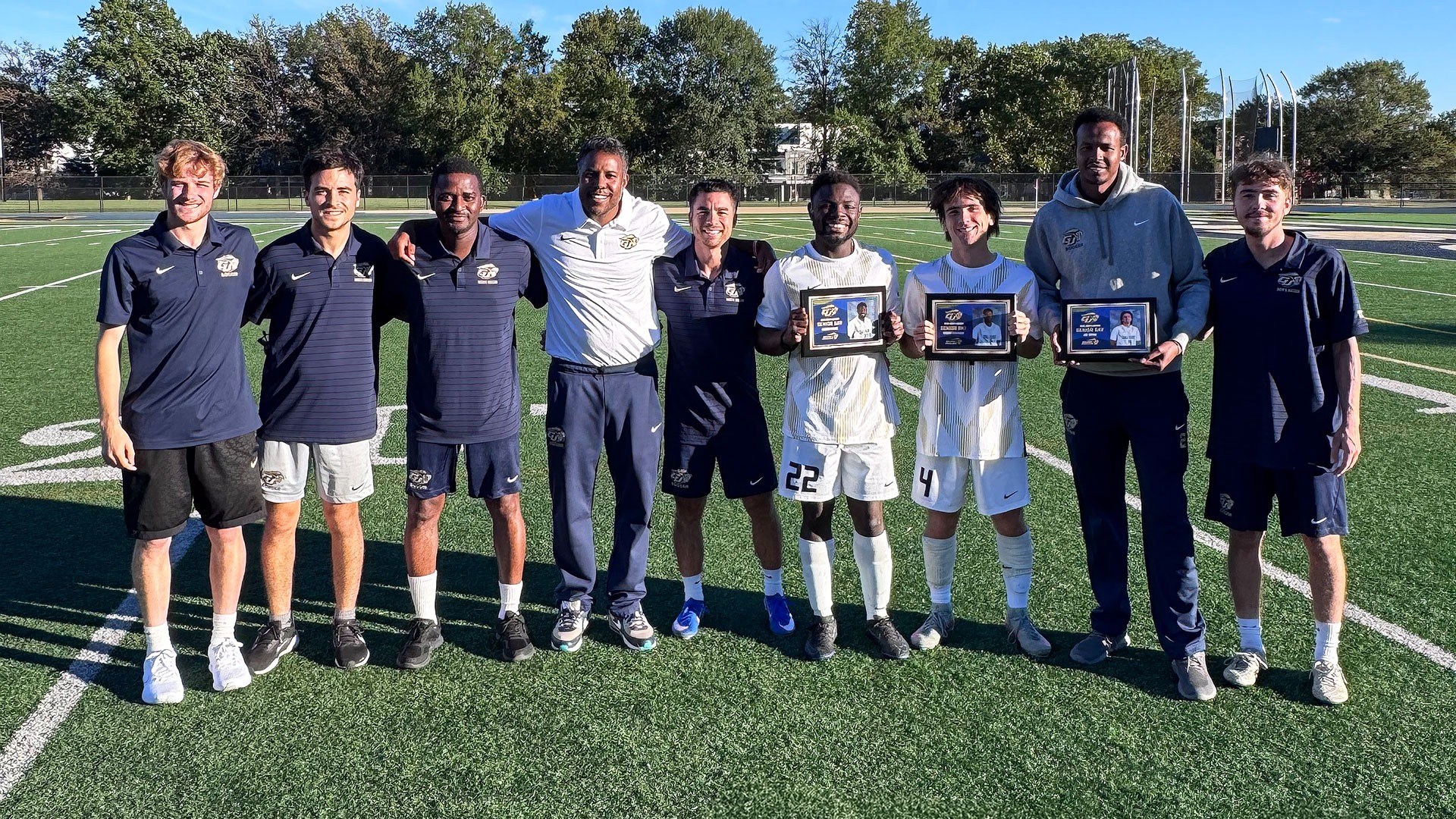 Gallaudet University men's soccer coaching staff pose alongside three players recognized during Senior Day on Oct. 21, 2026, at Hotchkiss Field. Gideon Nyame (#22), Joe Bryan (#4) and Sadam Mohamed (#2) stand together holding their framed Senior Day plaques, surrounded by six members of the Bison coaching staff. The group smiles for a photo on the field under sunny skies, with trees and a clear blue sky in the background.