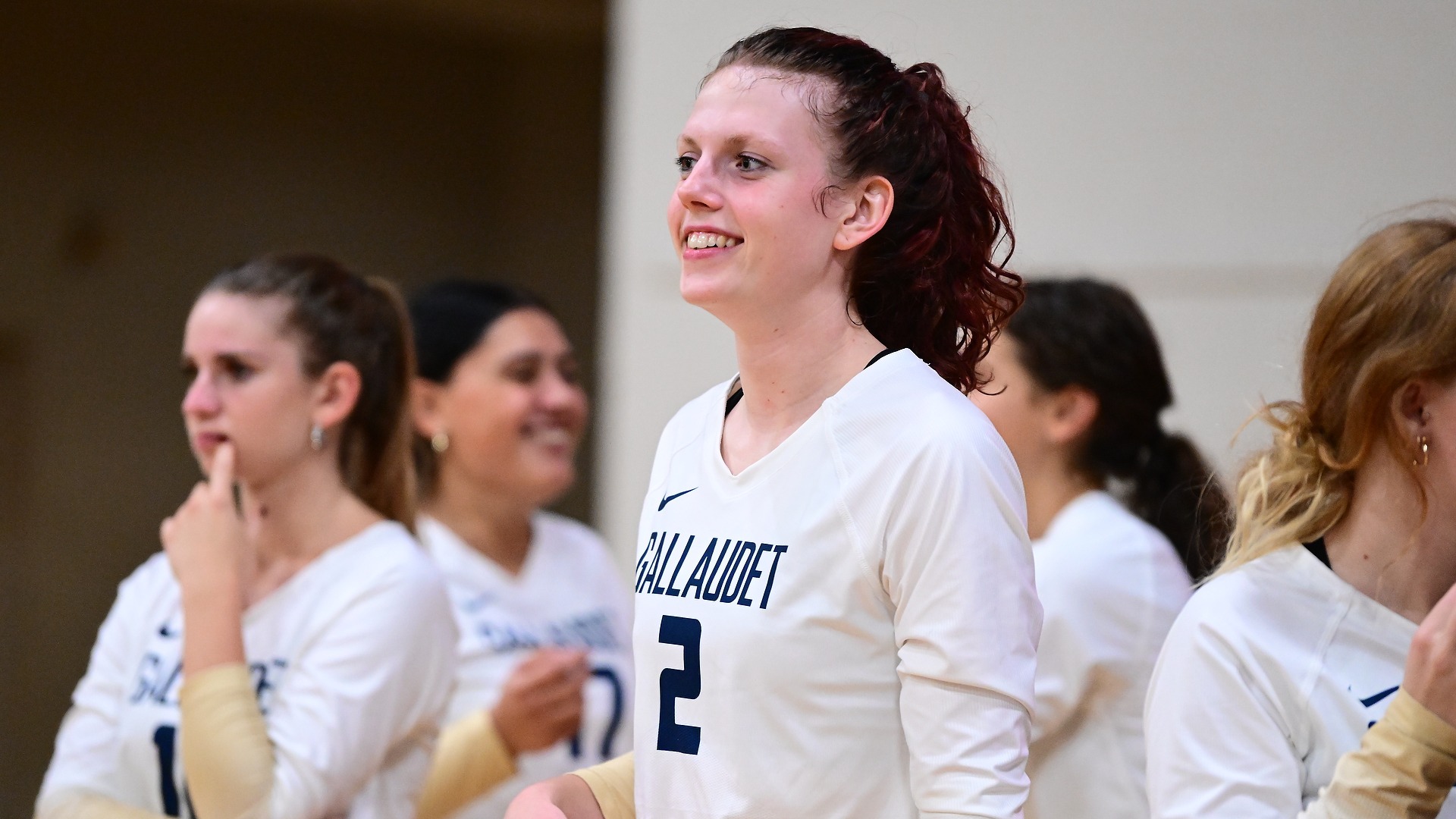 Gallaudet women's volleyball player Nirveli Smith (#2) smiles while standing on the sideline in her white-and-navy Bison uniform during a match, with several teammates gathered behind her.