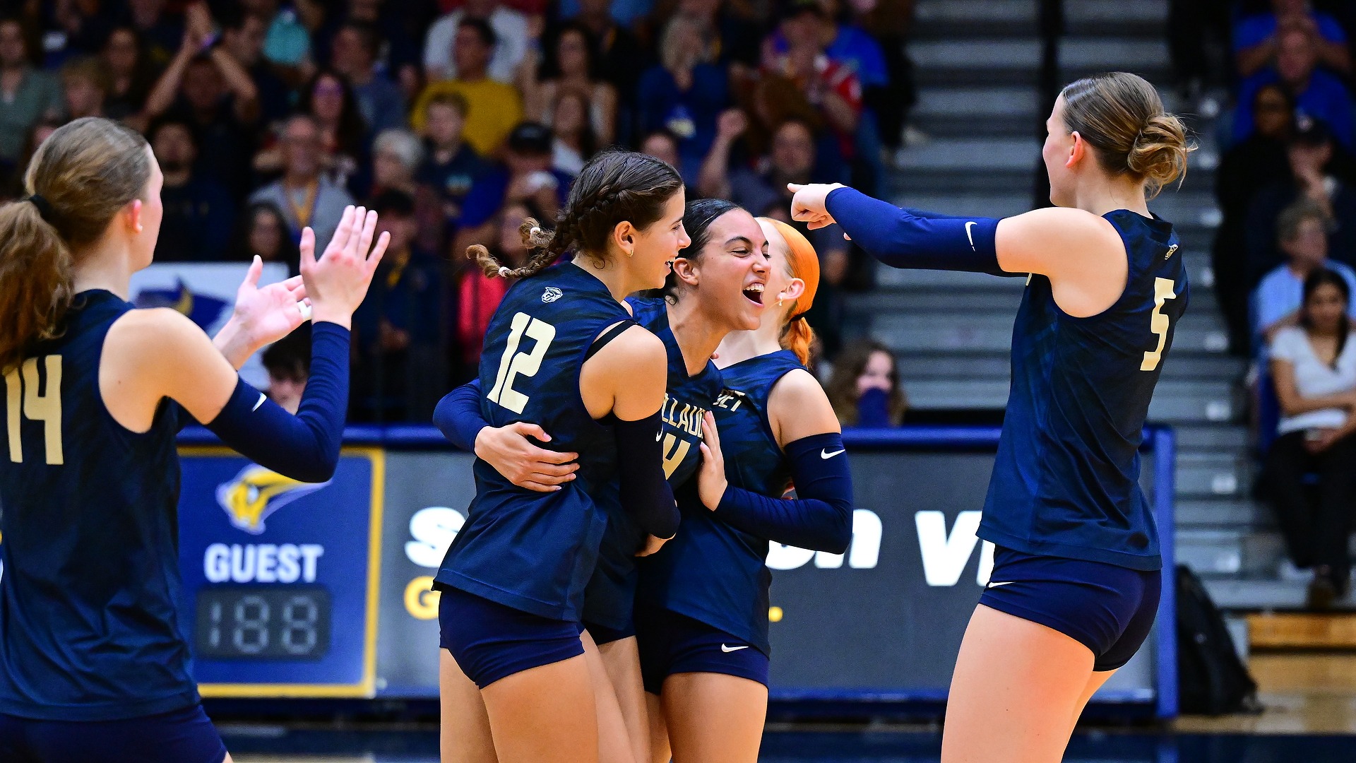Gallaudet women's volleyball player Gracie Gloster (#12) embraces teammate Bri Eilderts (#4) in celebration after the Bison secure a set victory, while Brielle Worley (#14) applauds on the left and Jada Zarembka (#5) points toward her teammates on the right during a match at the Field House