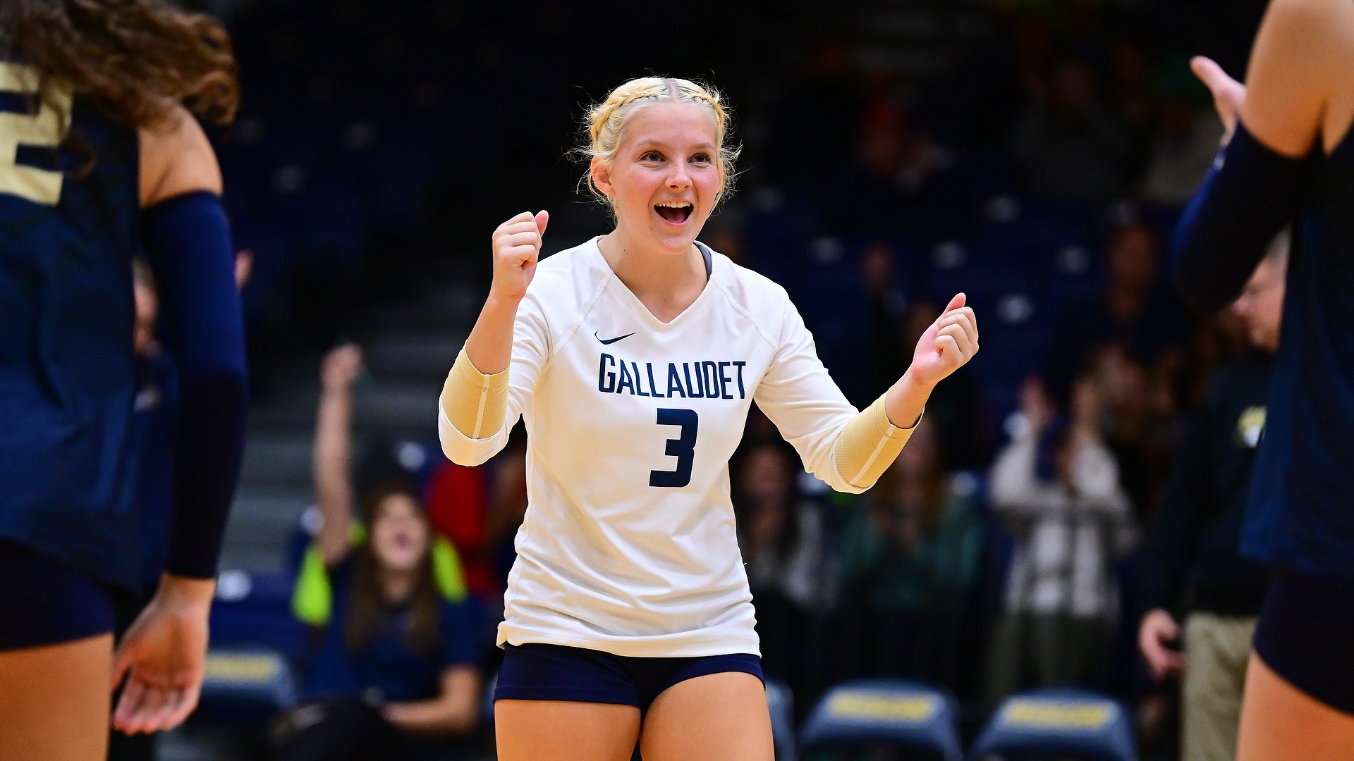 Gallaudet women’s volleyball player Sirrah Wilding (#3) celebrates a point with her fists raised and a smile as teammates surround her during a match in the Field House.