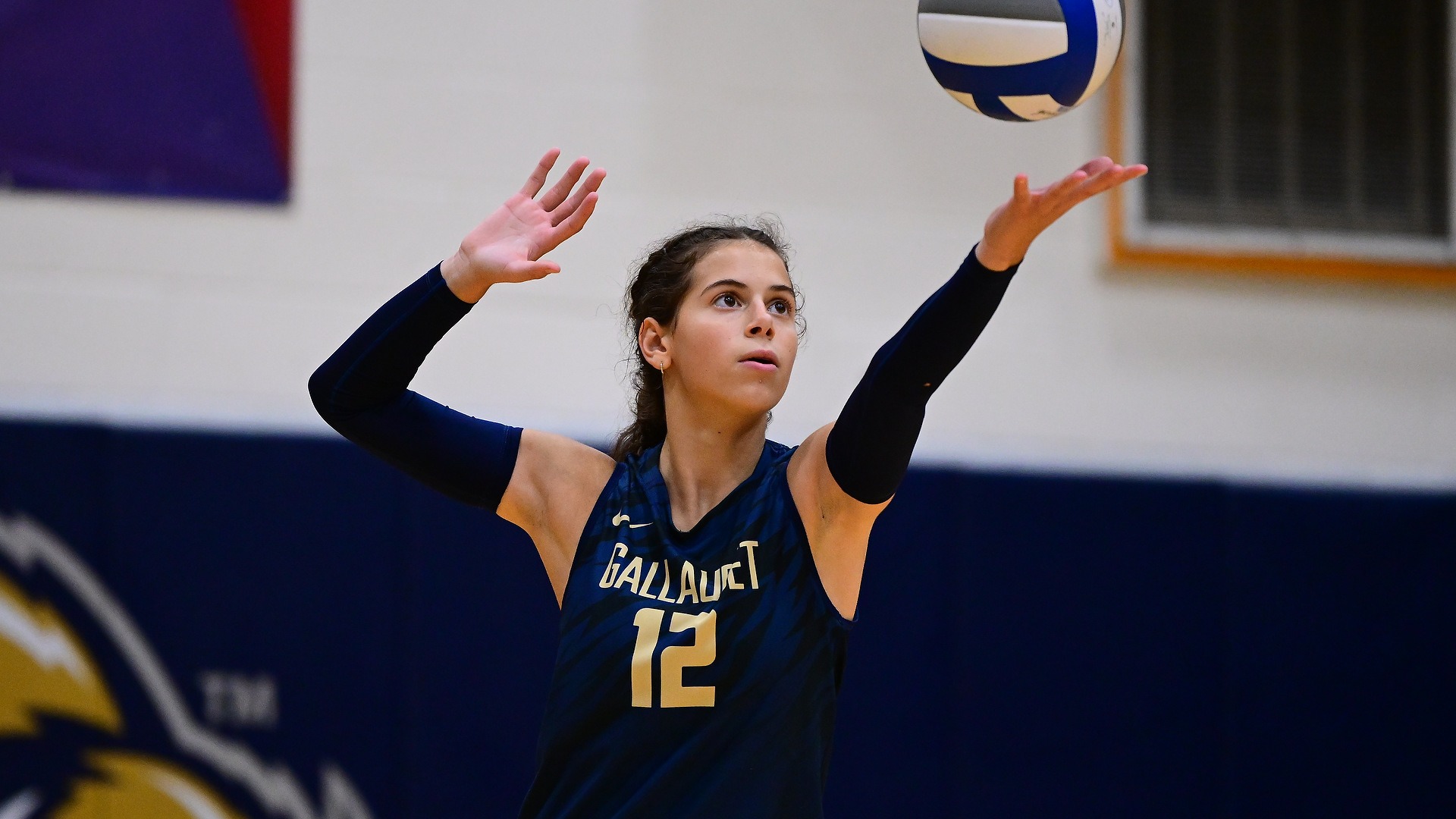 Gallaudet women's volleyball player Gracie Gloster (#12) tosses the ball into the air as she prepares to serve during a match.
