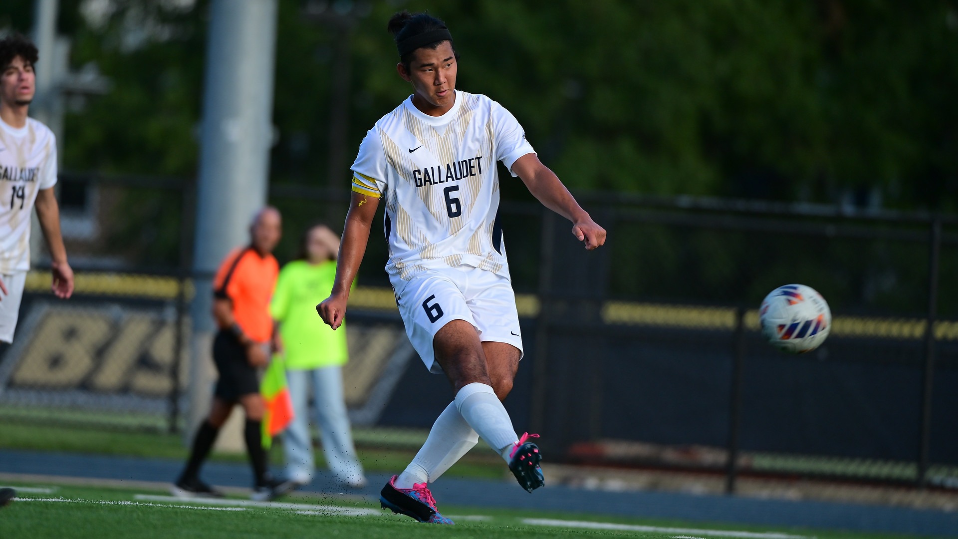Gallaudet men’s soccer player Eloi Straub (#6) kicks the ball downfield in his white-and-gold Bison uniform during a match at Hotchkiss Field, with a teammate and sideline officials in the background.