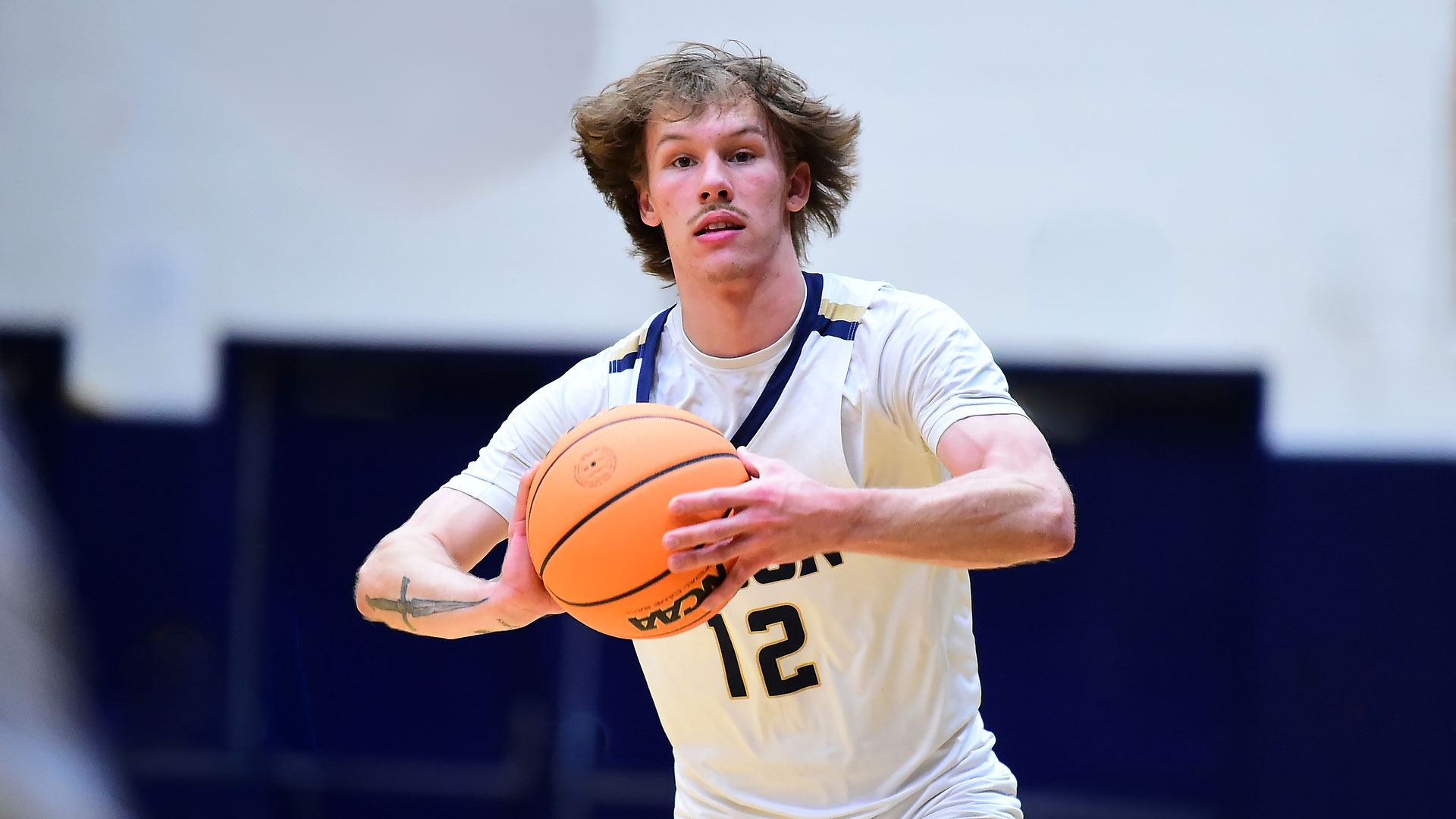 Gallaudet University men's basketball player Alex Ortman gets set to make a pass to a teammate during a game in the Field House. The GU player is wearing an all white uniform with the word BISON across his chest in Navy blue font and the #12 in navy blue.