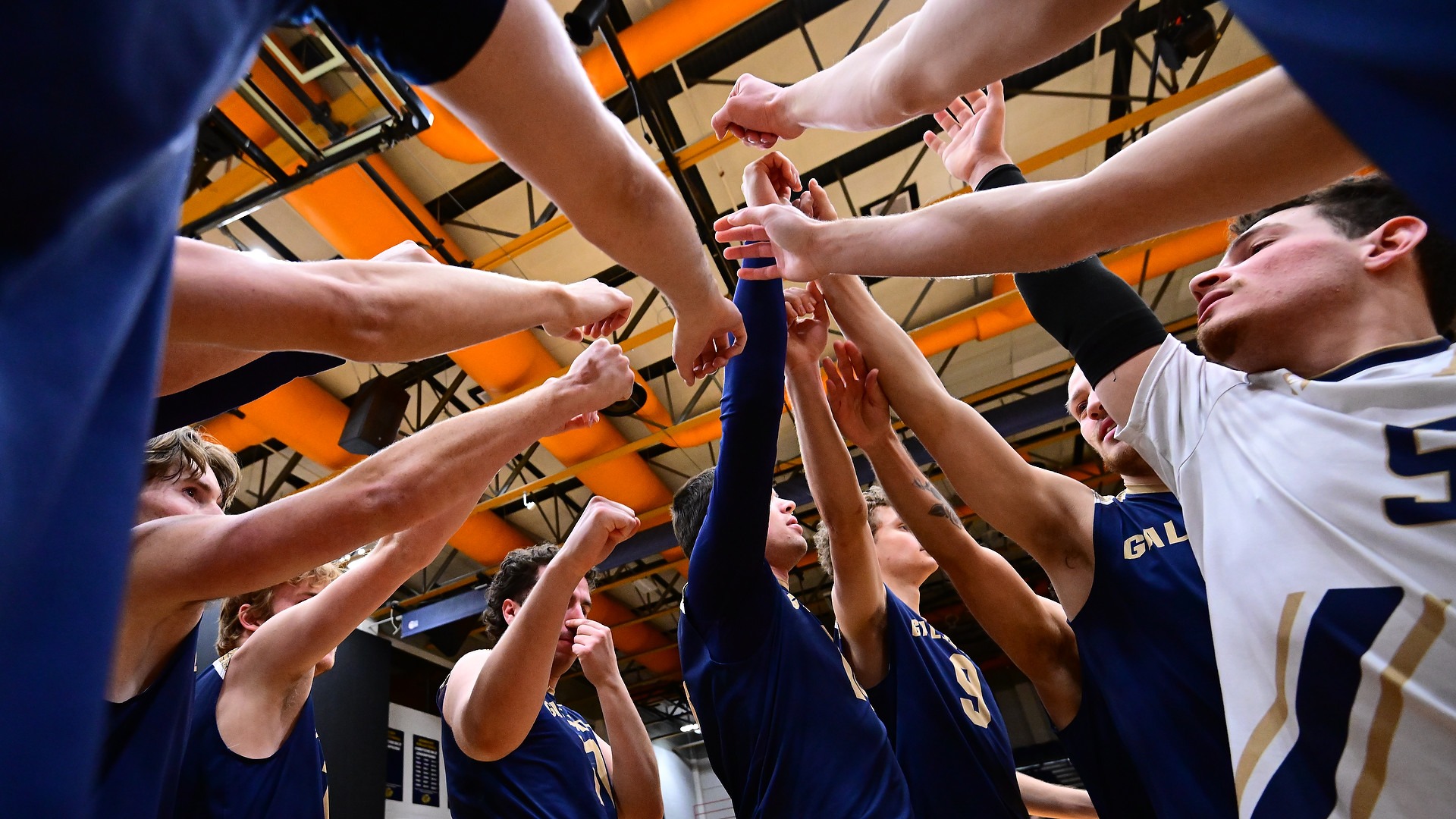 Gallaudet men's volleyball team huddles up before a match in the Field House. Each player raises one of their arms to the sky together before they break the huddle.