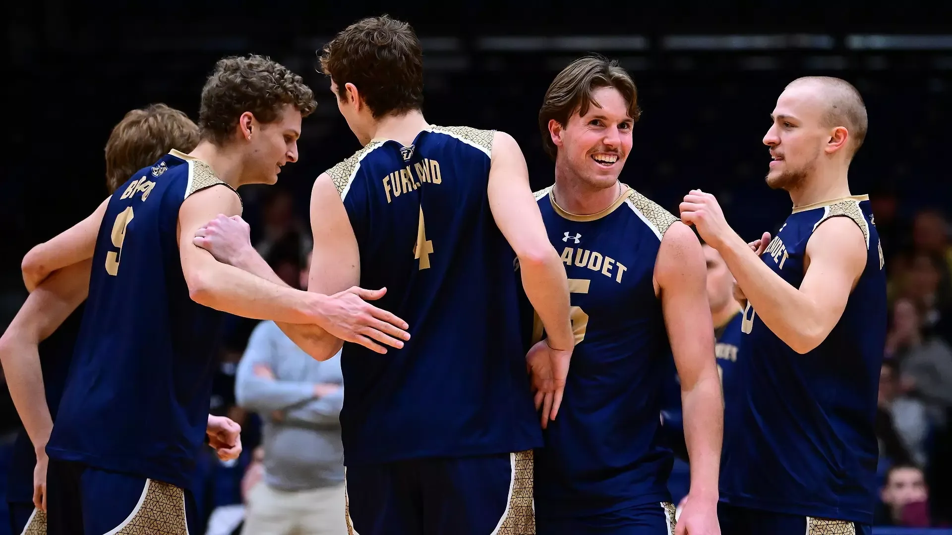 Gallaudet men's volleyball team celebrates a point during a home match in the Field House. The six players come together to huddle up and are smiling at each other in excitement. Sophomore Kodi Lee smiles and looks toward the camera.
