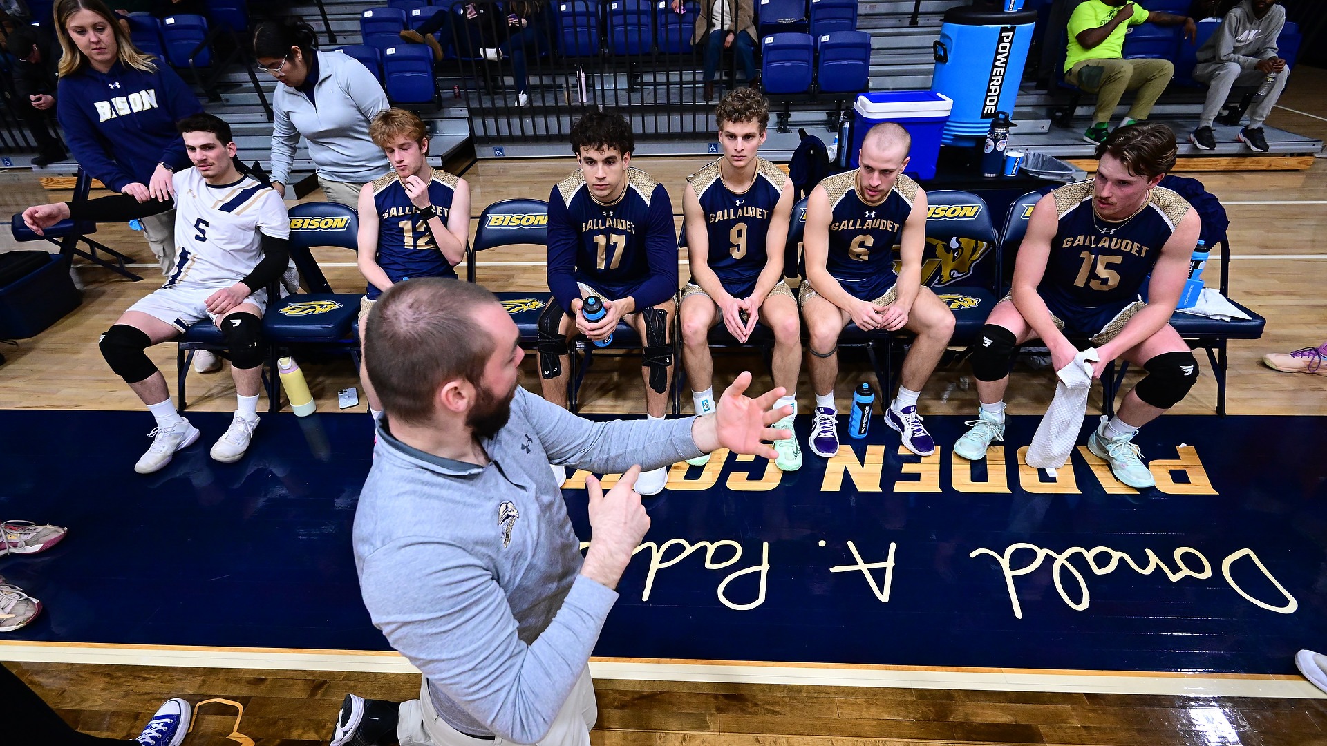 Gallaudet men's volleyball coach Ben Campman signs with his team during a timeout as the Bison players sit on the bench during the break.
