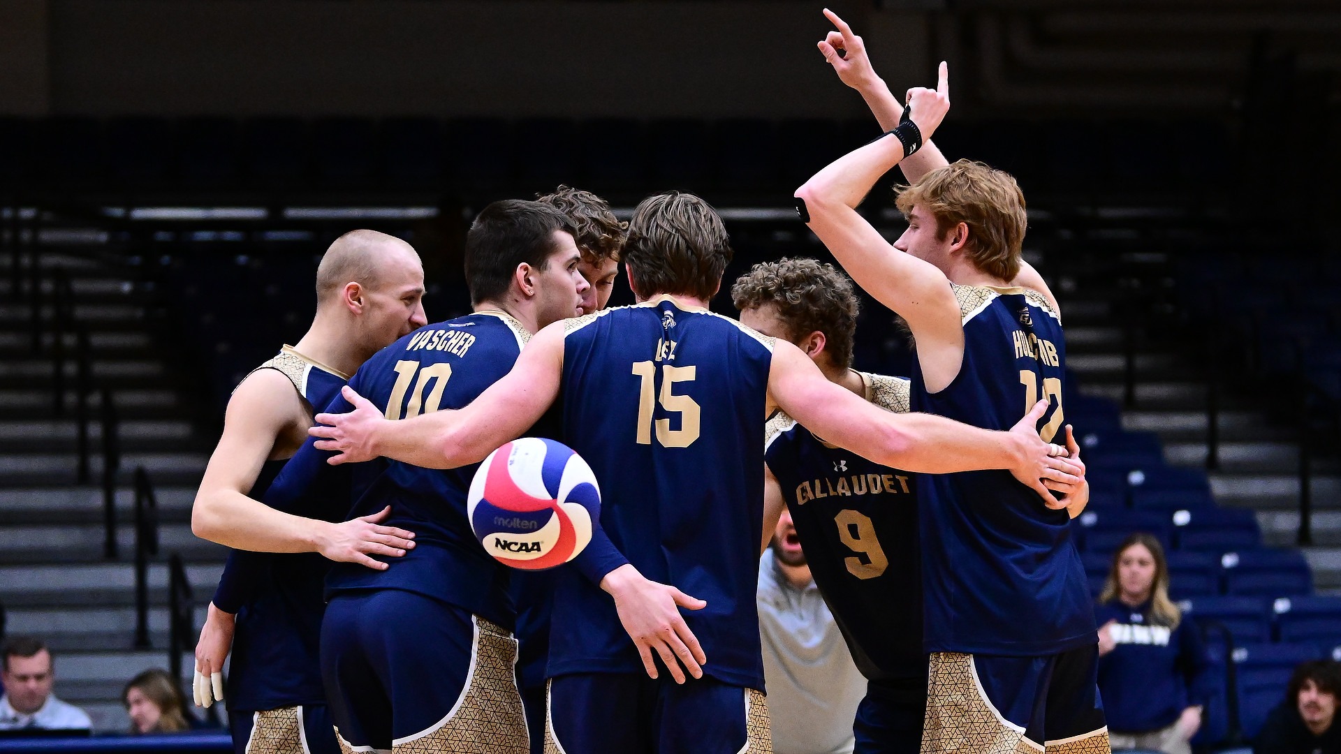 Gallaudet men's volleyball team celebrates a point during a home match in the Field House. The six players come together to huddle up and are smiling at each other in excitement.