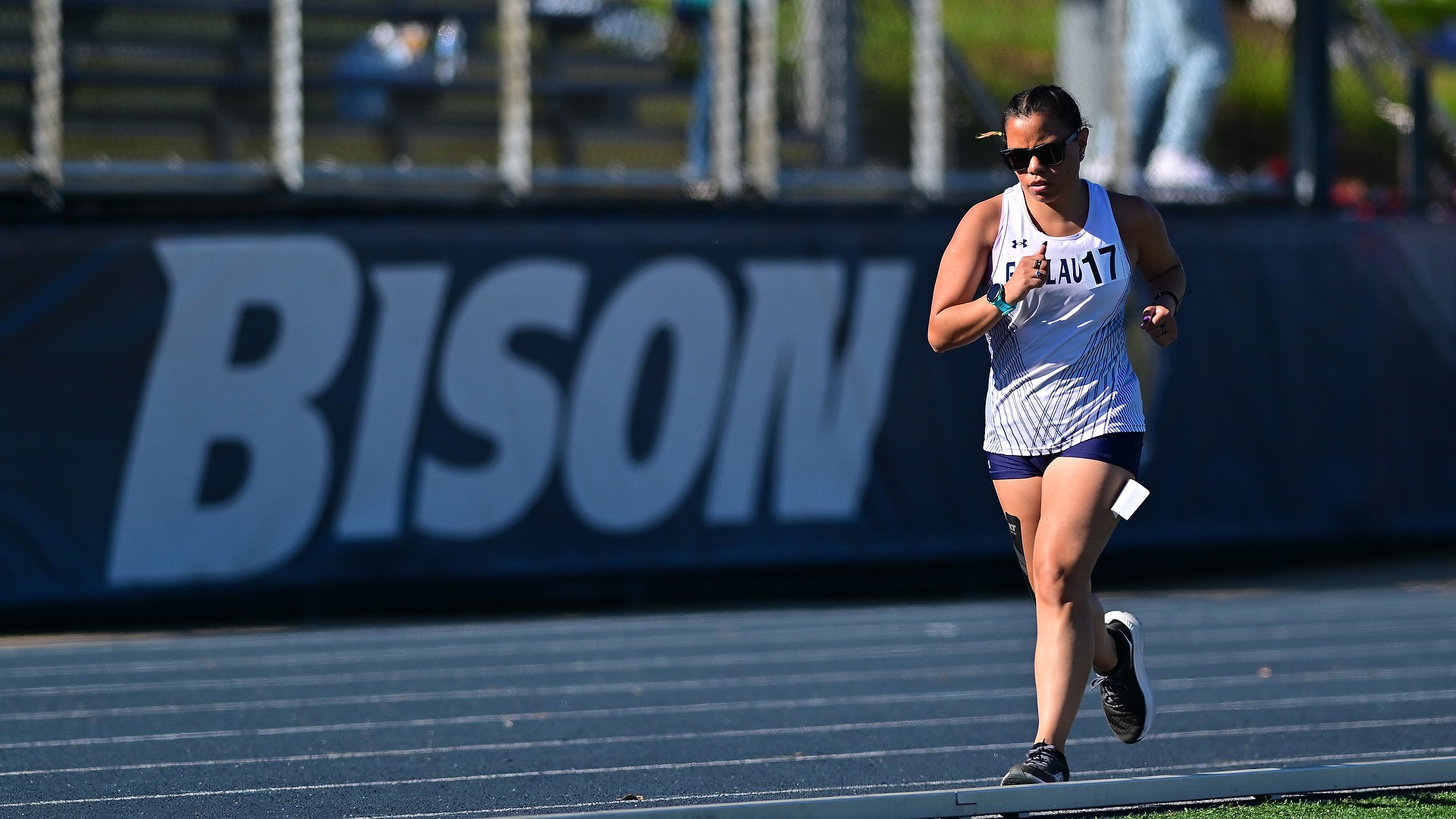 Gallaudet women's track and field long-distance runner Olivia Armstrong runs on the blue track on the campus of Gallaudet University as the afternoon sun begins to set. The word BISON is in large white font on a banner attached to the grandstand behind Armstrong.