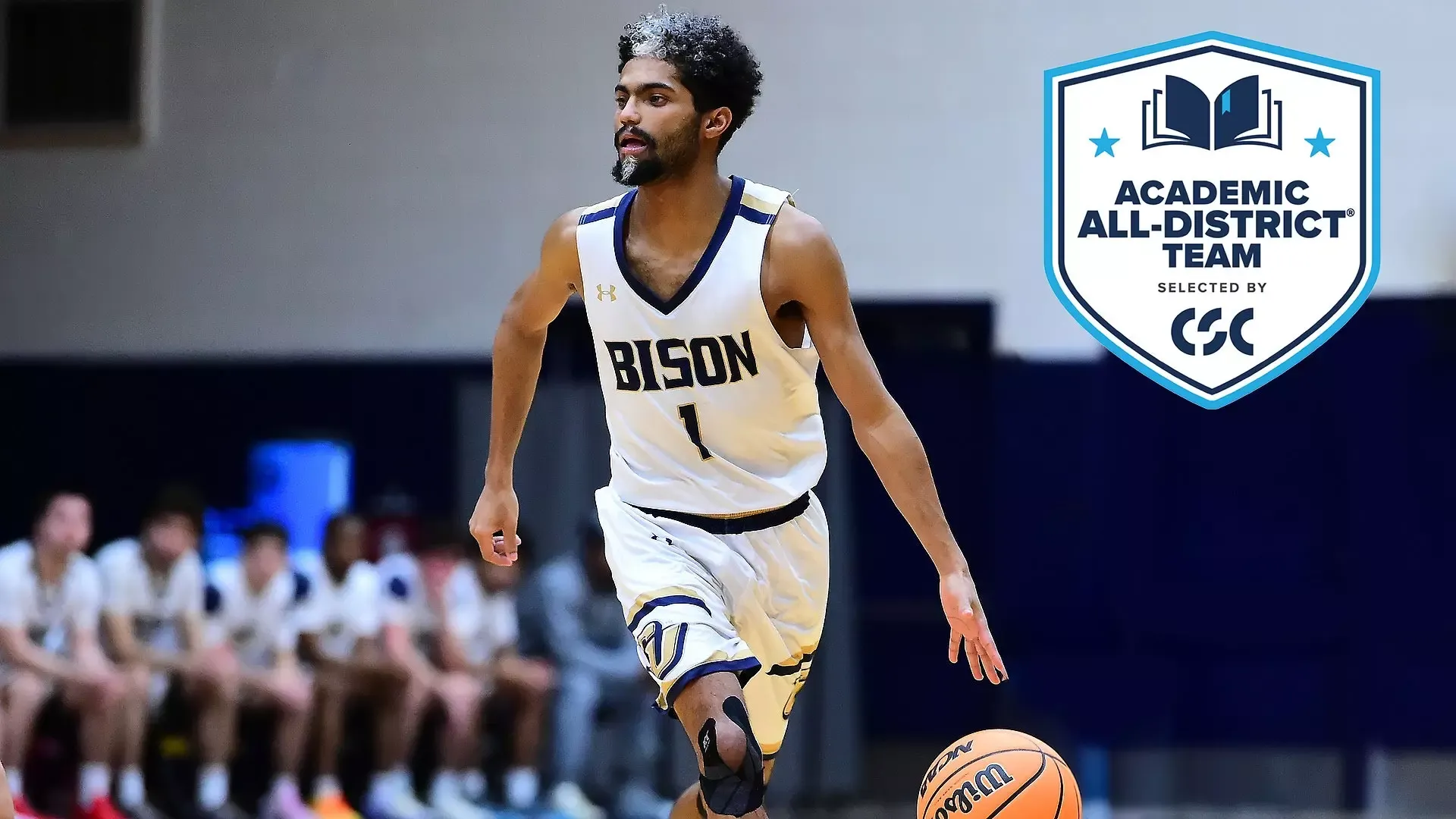 Gallaudet men's basketball player Bruce Brewer Jr. dribbles a basketball during a home game in the Field House. In the upper righthand corner is a College Sports Communicators Academic All-District logo in the shape of a shield. 