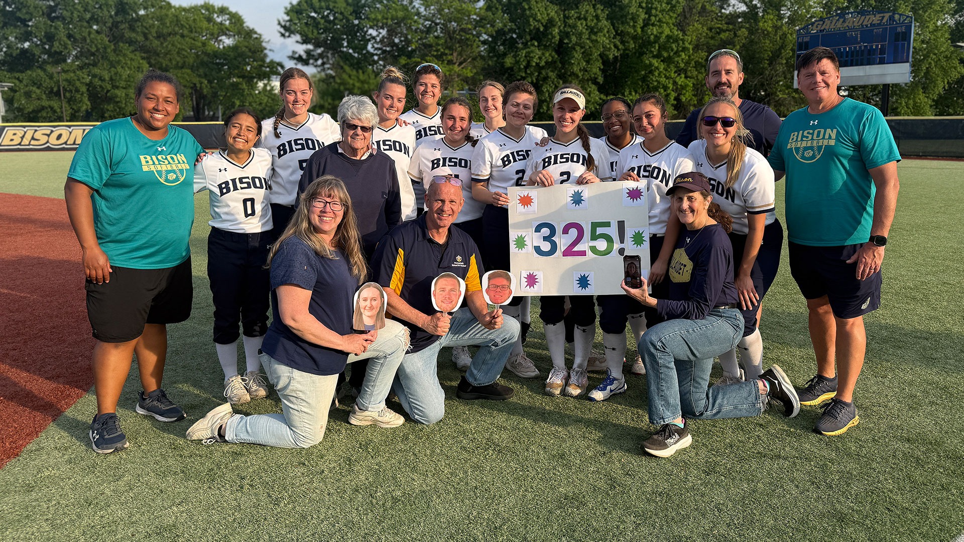 The Gallaudet softball team gathers around senior pitcher Lacey Drolsbaugh and the Drolsbaugh family members in attendance to witness Lacey's career setting game as she became the all-time strikeout leader for the Bison with 325. Lacey holds up a poster with the numbers 325 on it as the sun sets in the distance.