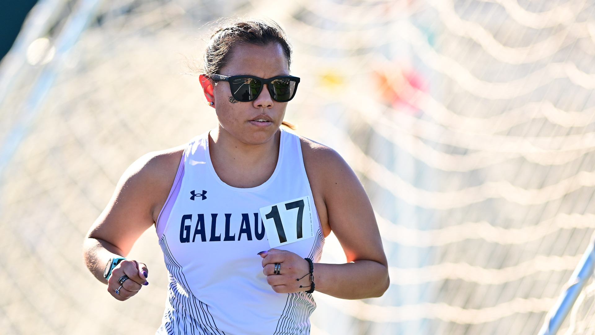 Gallaudet women's long-distancer runner Olivia Armstrong runs in an event on a sunny afternoon. Armstrong wears black sunglasses and a white Gallaudet tank top uniform. 