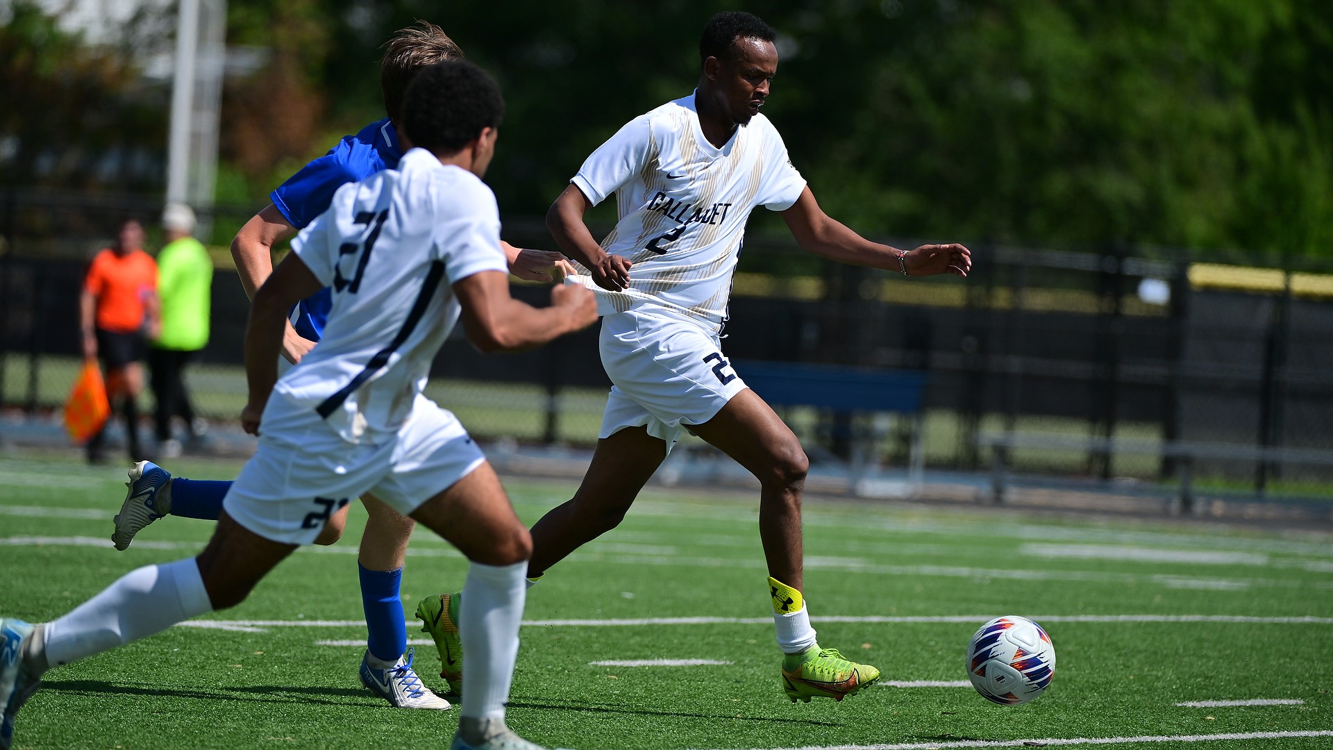 Gallaudet men’s soccer player Sadam Mohamed (#2) dribbles the ball upfield in his white Bison uniform while being held by an opposing defender during a match on a sunny afternoon.