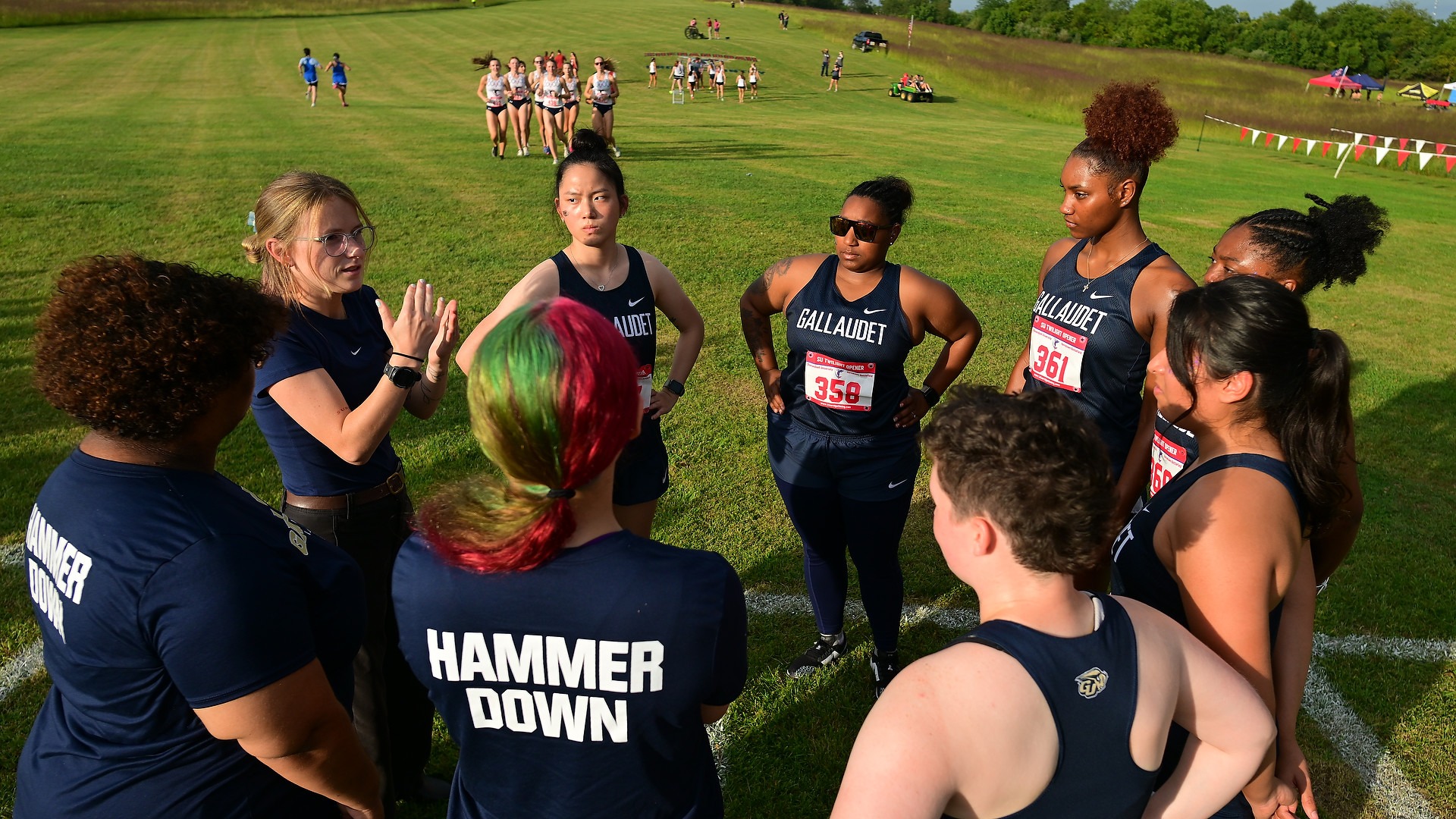 Gallaudet cross country Associate Head Coach Nykol Pratt signs a pre-race pep talk to the Bison women’s team gathered in a huddle on a grassy field at the SU Twilight Open in Winchester, Virginia, as the soft light of the setting sun falls across the team.