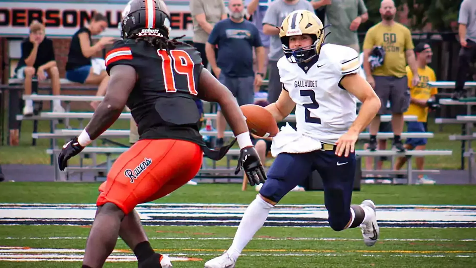 Gallaudet quarterback Jayden DeFalco (#2), wearing a white Bison jersey, navy pants and a gold helmet, carries the football toward an Anderson University defender (#19) dressed in a black jersey, orange pants and black helmet during Saturday’s game in Anderson, Indiana.