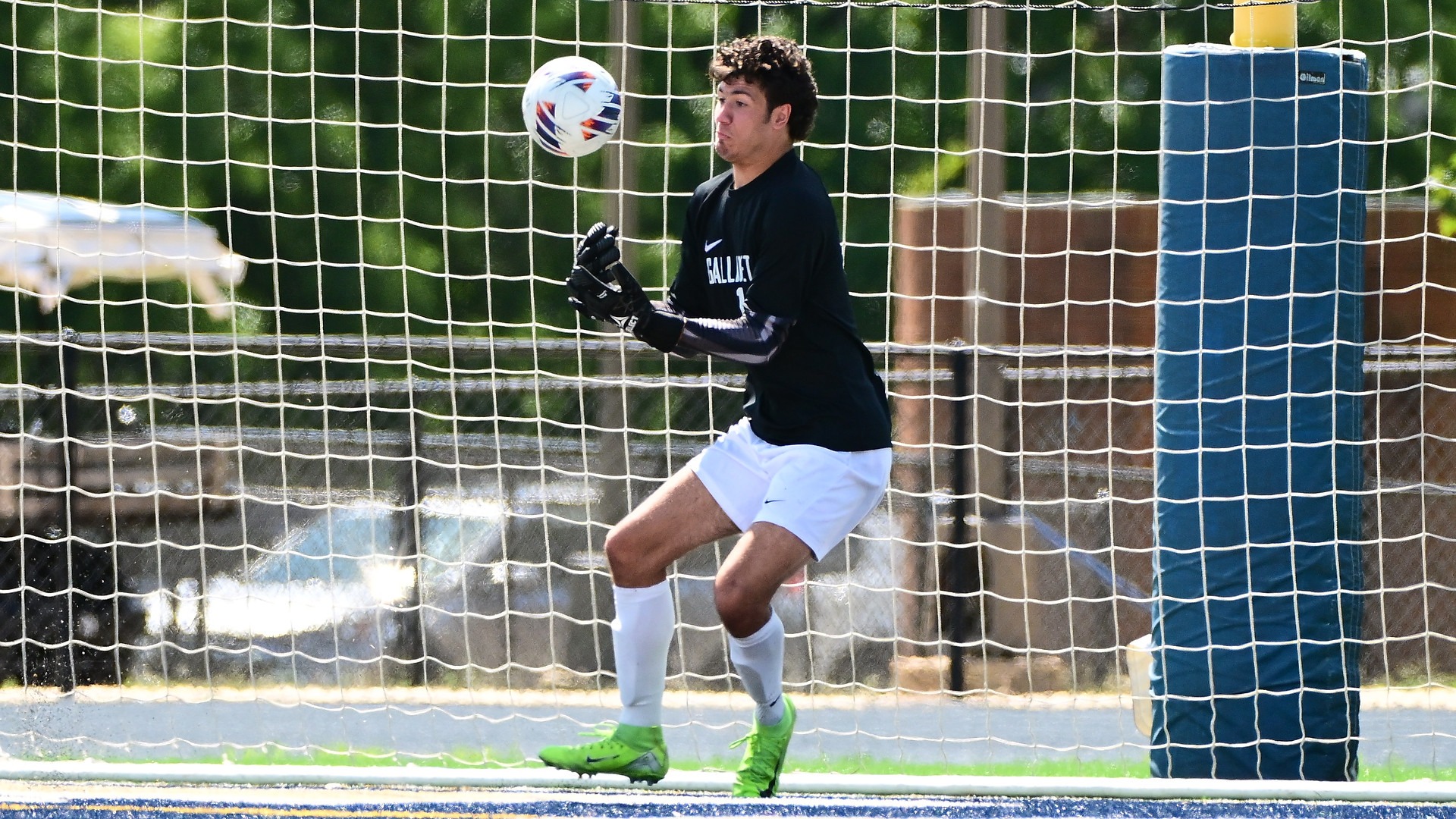 Gallaudet goalkeeper John McCabe makes a save in front of the net during a men's soccer game on a sunny day at Hotchkiss Field. McCabe wears a black goalkeeper jersey, white shorts, white socks, black gloves and bright green cleats as he tracks the ball in front of the goal.