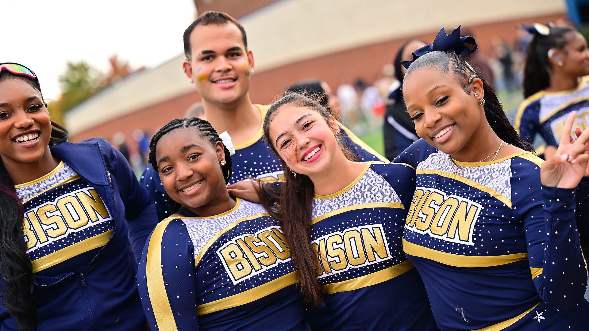 Gallaudet cheerleaders smile and pose together on the sidelines in navy, gold and white Bison uniforms during a football game at Hotchkiss Field.