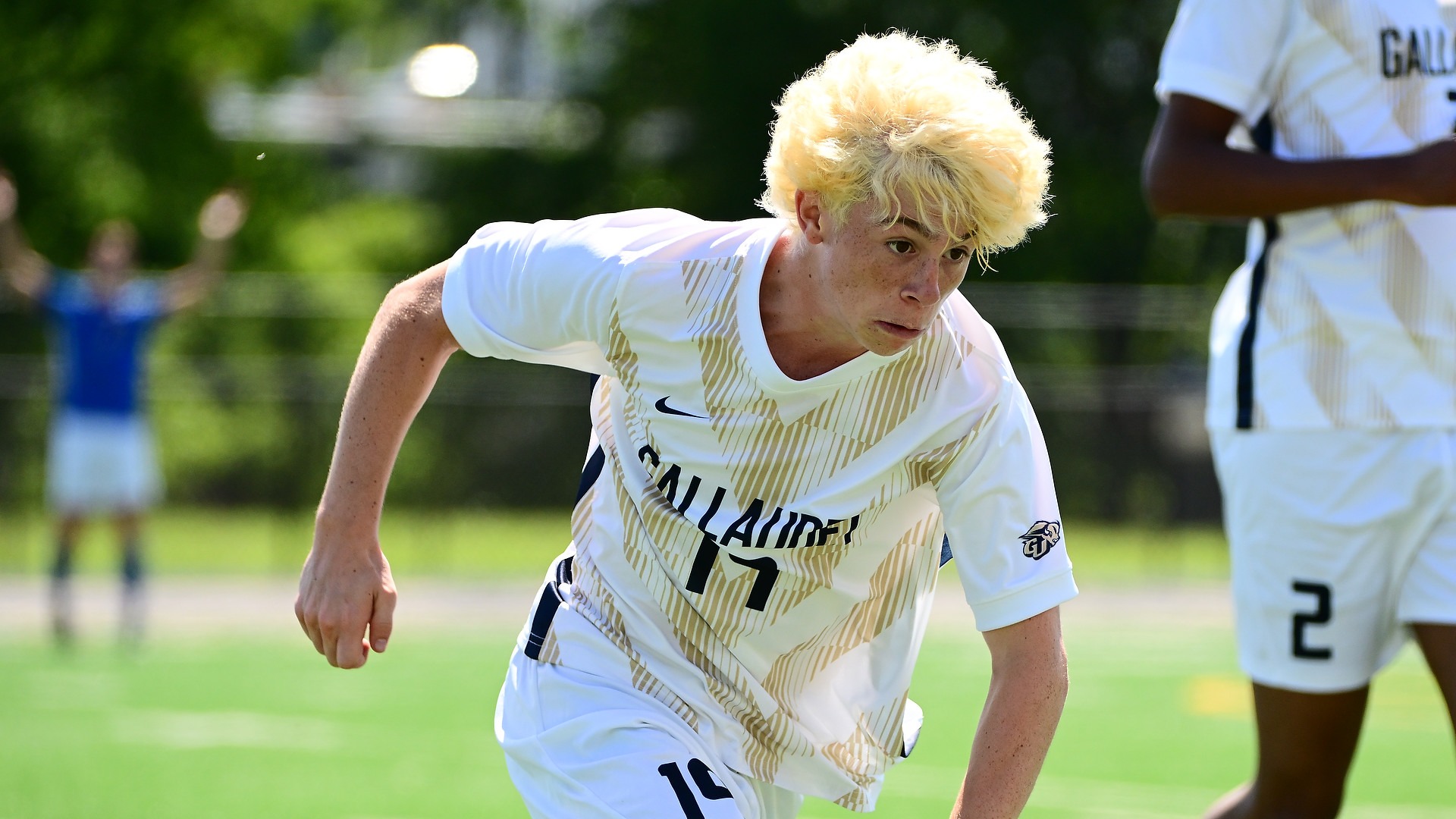 Gallaudet men’s soccer player Eli Matamoros (#19), with bright blond hair, leans forward in action while wearing the Bison’s white uniform with gold geometric accents on a sunny afternoon. The green field, trees, spectators and a Gallaudet teammate are softly blurred in the background.
