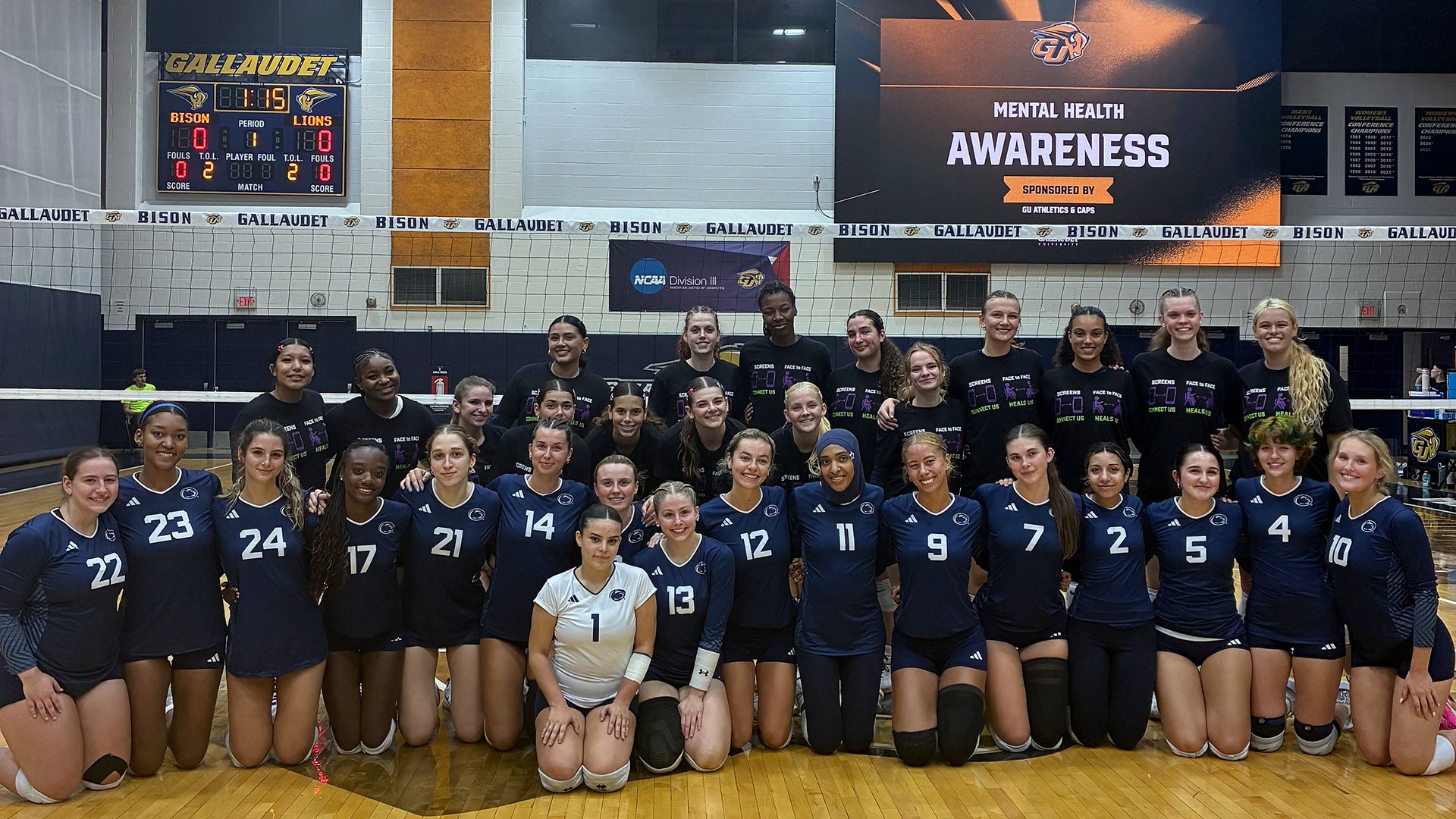 The Gallaudet and Penn St.-Abington women’s volleyball teams come together for a group photo on the court before their match at the Field House as part of Mental Health Awareness Night. Players wear their team uniforms or black mental health awareness shirts, with the videoboard displaying “Mental Health Awareness” above the group.