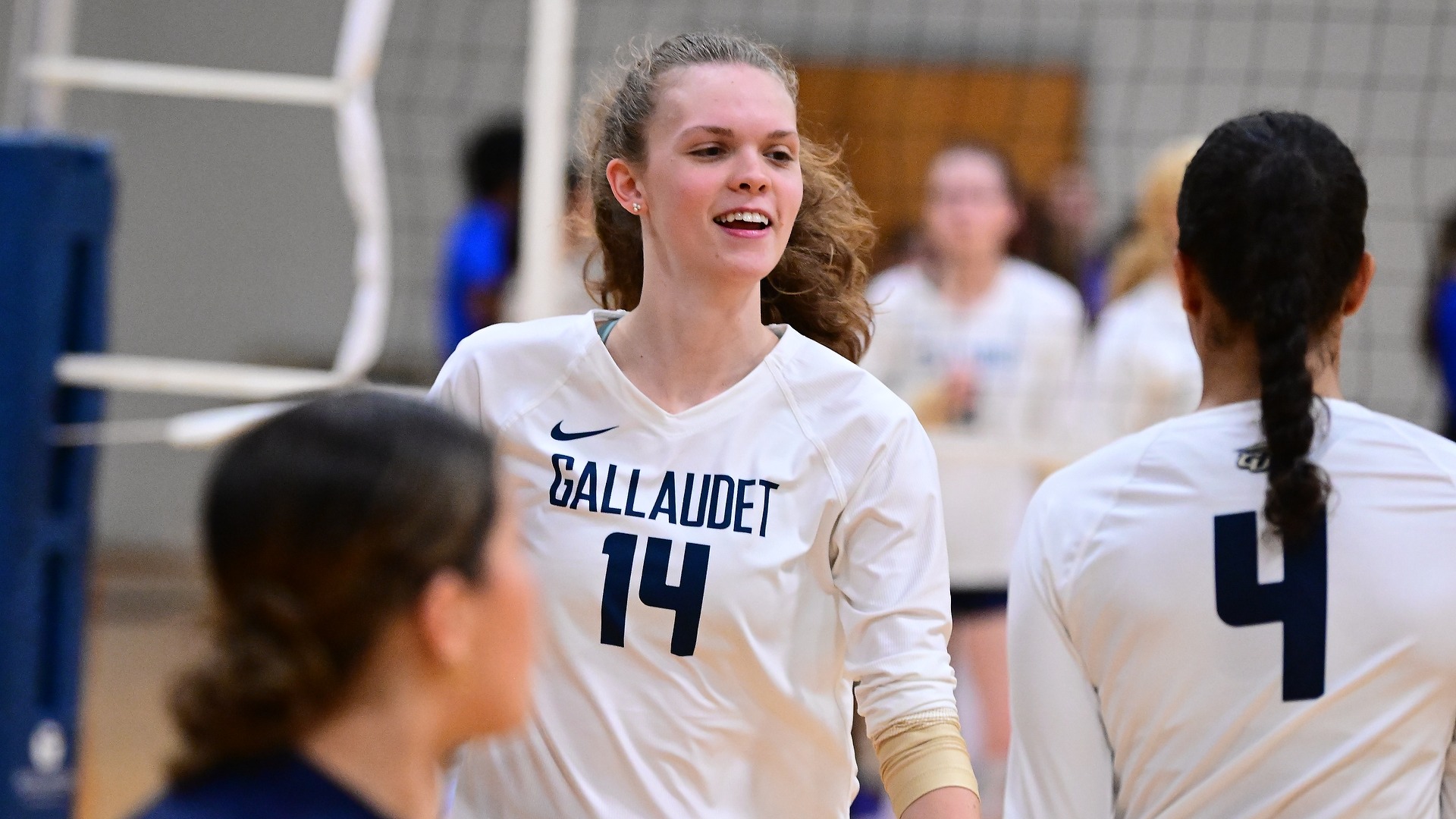 Gallaudet women’s volleyball player Brielle Worley (#14) smiles while wearing the Bison’s white long-sleeve jersey with navy lettering and numbers during warmups on the court, with teammates surrounding her.