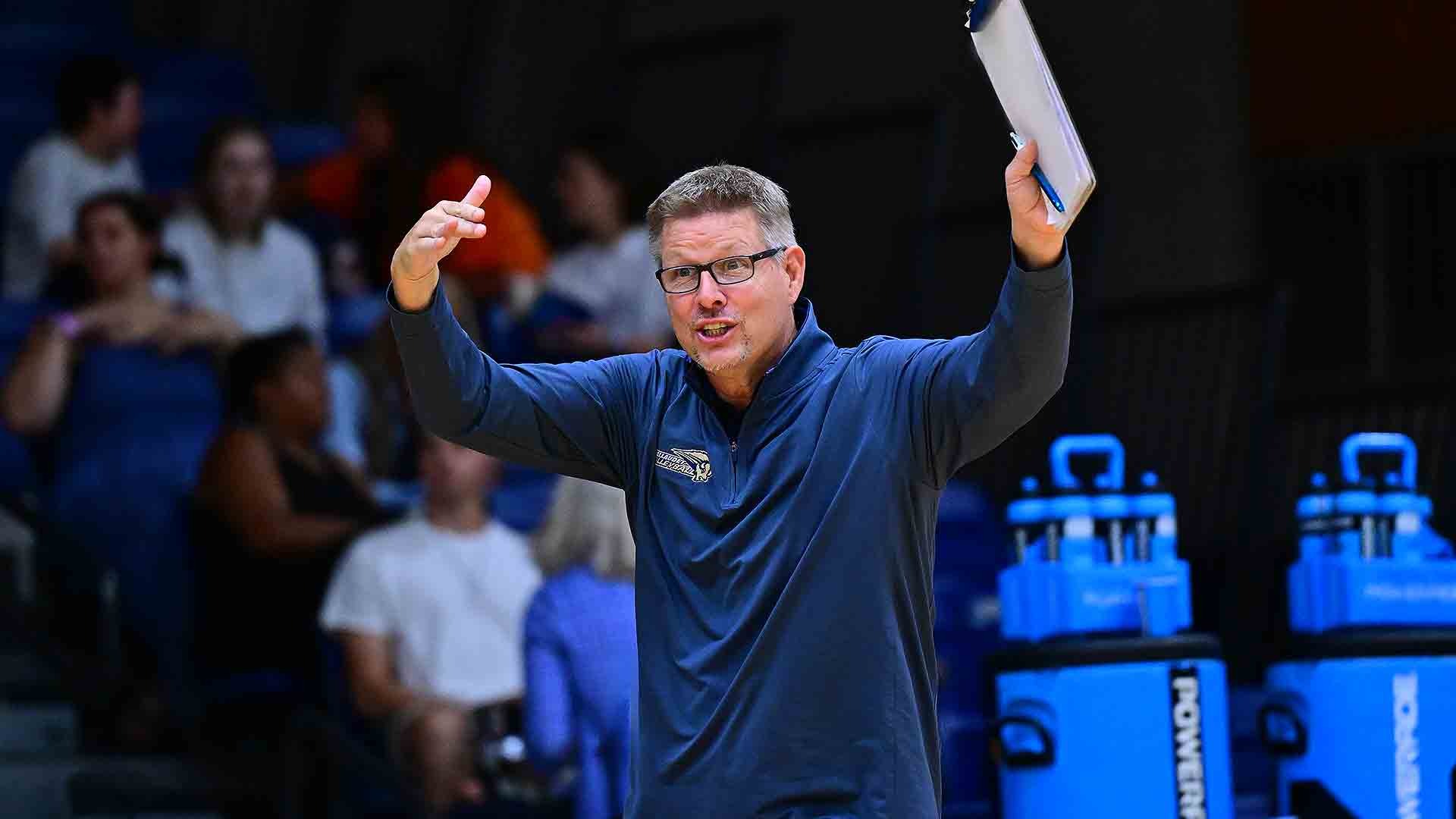 Gallaudet women’s volleyball head coach Lynn Ray Boren gestures with both arms raised while holding a clipboard on the sideline during a match.