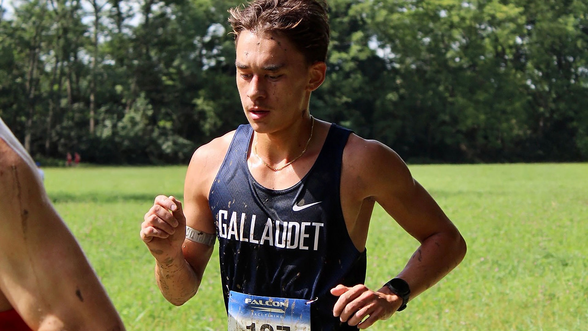 Gallaudet sophomore Sevan Ikeda runs during the Dutchmen Invitational on a sunny and hot Saturday morning. Ikeda is shown close up in his Gallaudet navy uniform made by NIKE, sweating with mud and debris on his arms and jersey.