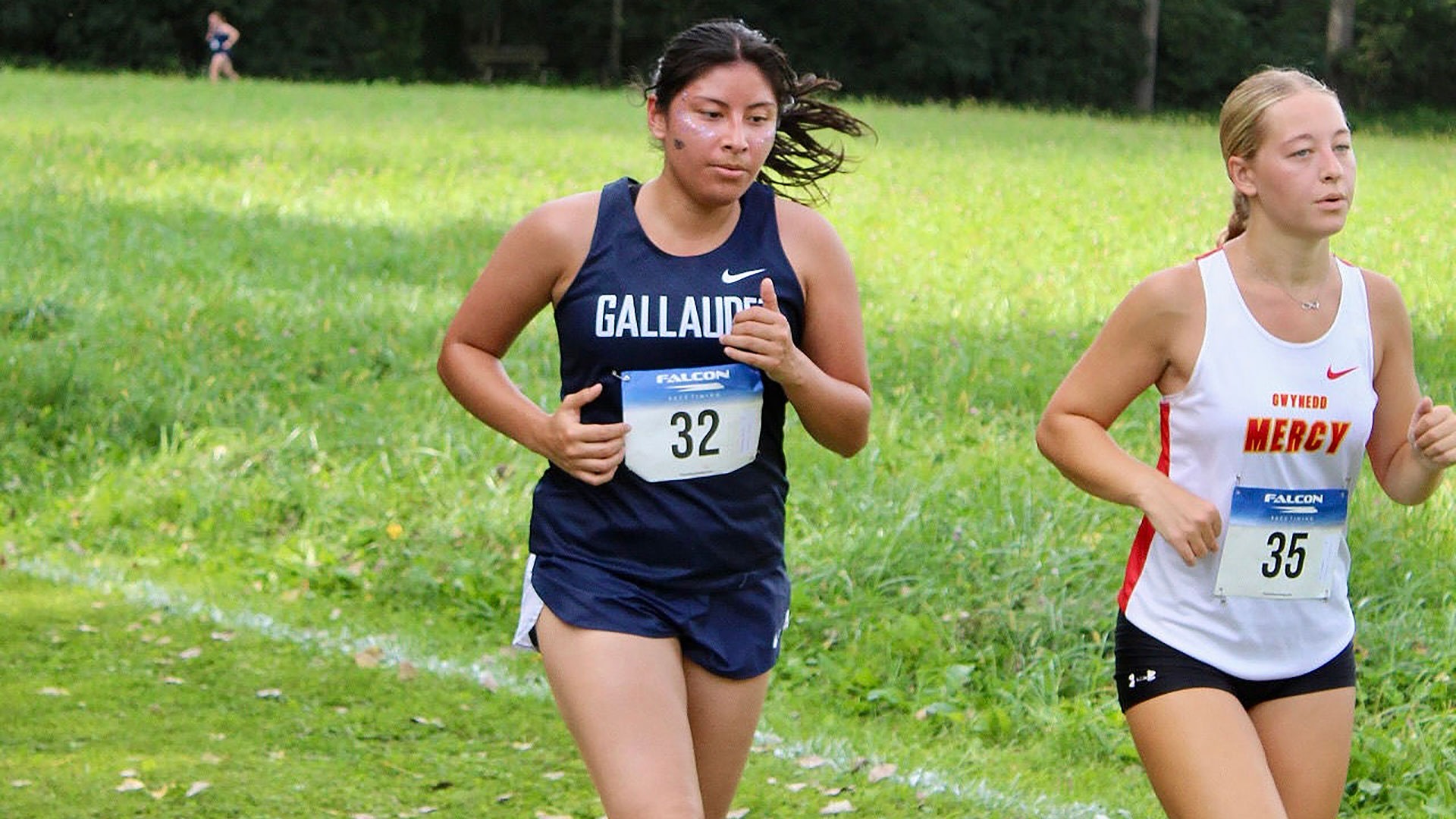 Gallaudet runner Ashley Morales-Juan competes at the Dutchmen Invitational on Saturday morning. Morales-Juan, wearing a navy colored Gallaudet uniform made by NIKE, runs through a shaded section of the course alongside another competitor.