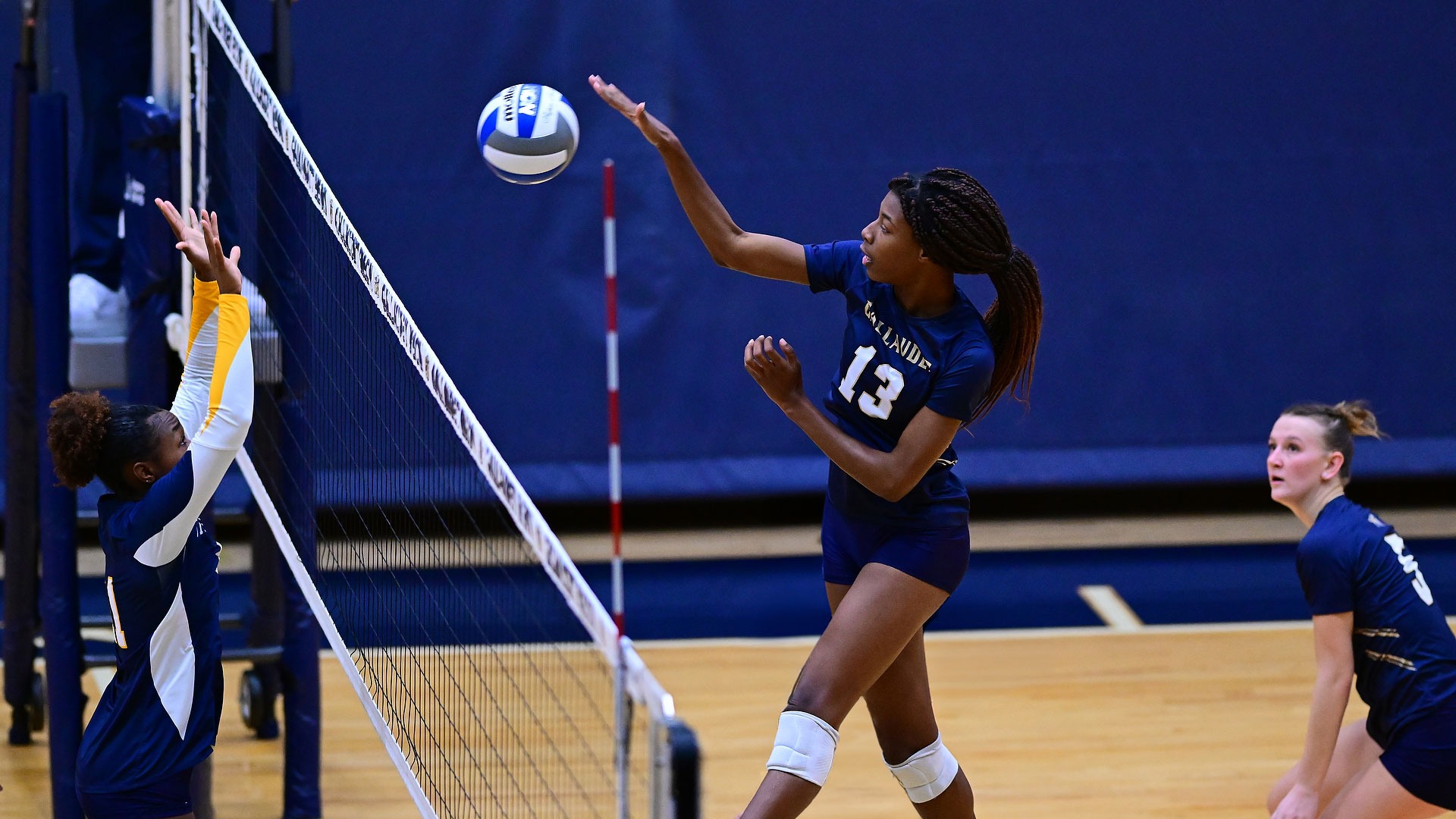 Gallaudet women’s volleyball player Kayla Debrow (#13) rises at the net and reaches high with her right arm to attack the ball as an opposing blocker attempts to defend during a match. A Gallaudet teammate looks on from the back row.