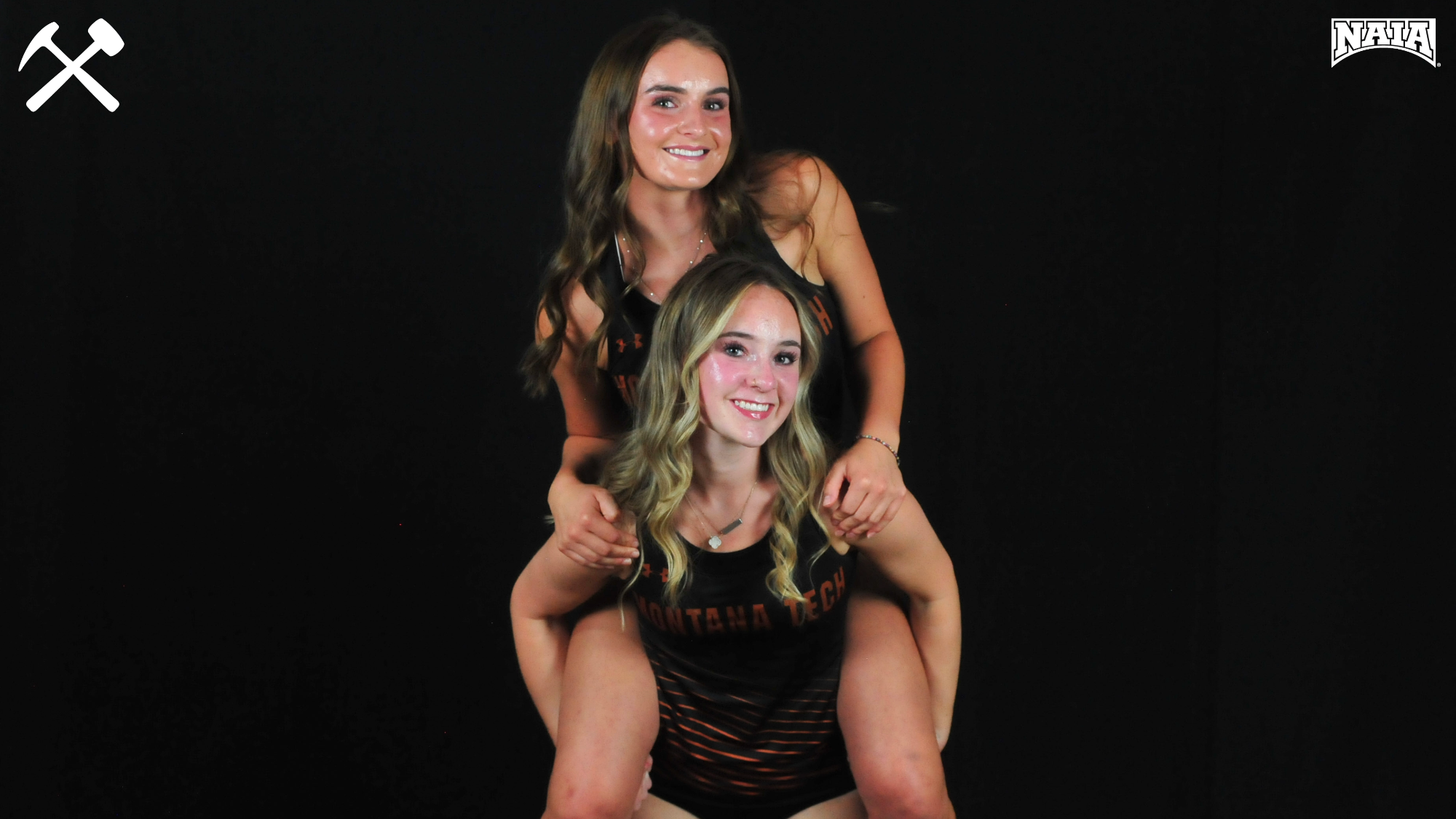 Montana Tech cross country runners Bella Bermingham and Hadyn Garza pose piggyback style in uniforms on a black background