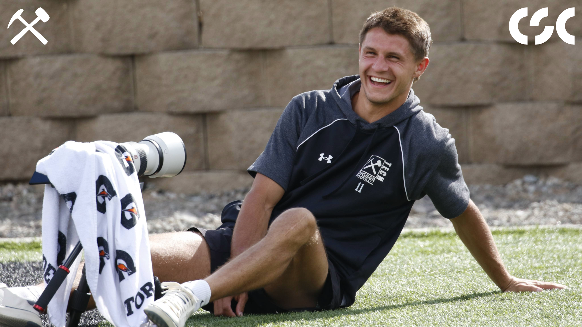 Kyle Torgerson smiles sitting behind his camera sitting on the turf at Alumni Coliseum
