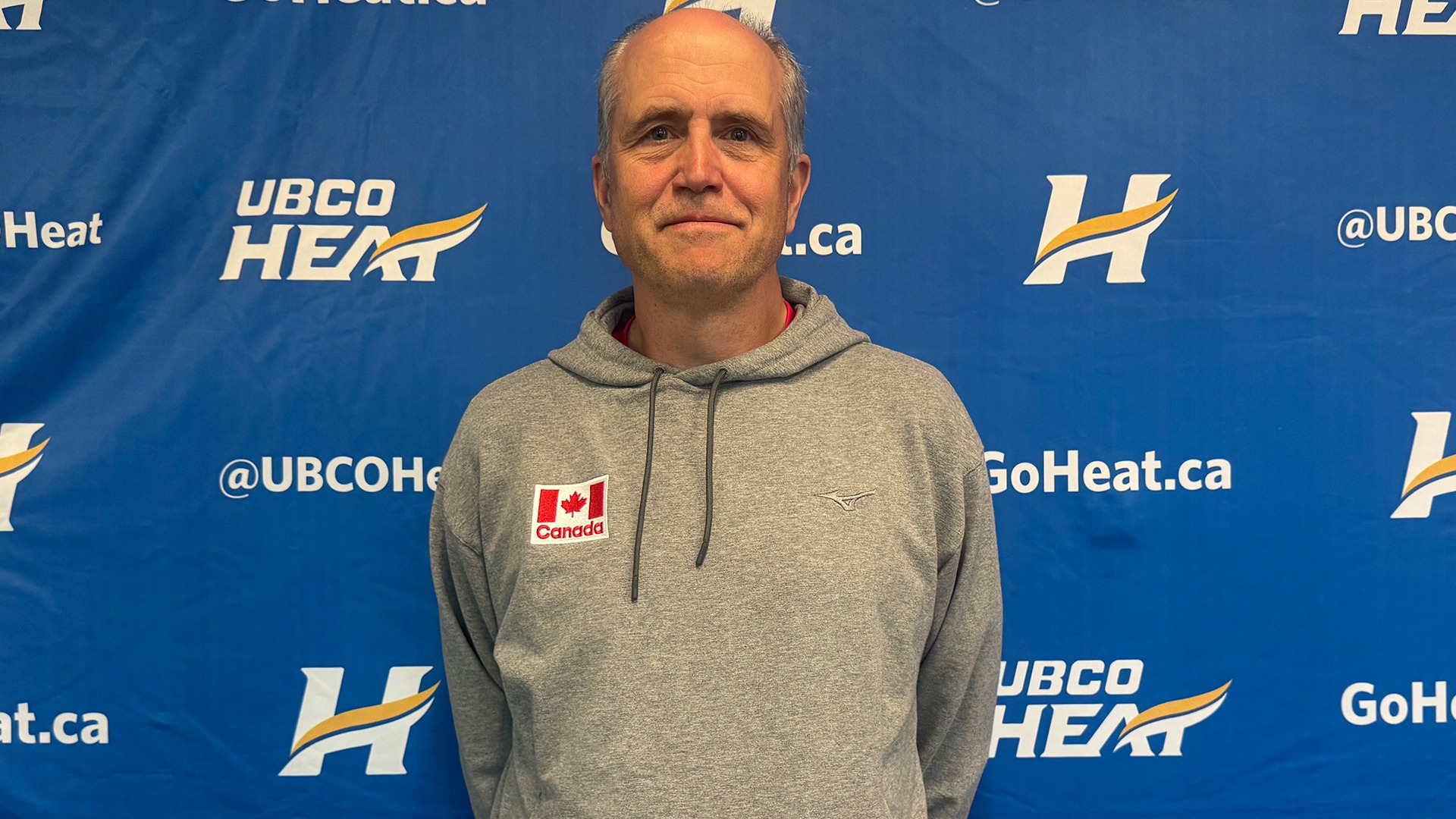 UBCO Heat men's volleyball head coach Scott Koskie stands in front of a UBCO Heat backdrop wearing Team Canada gear.