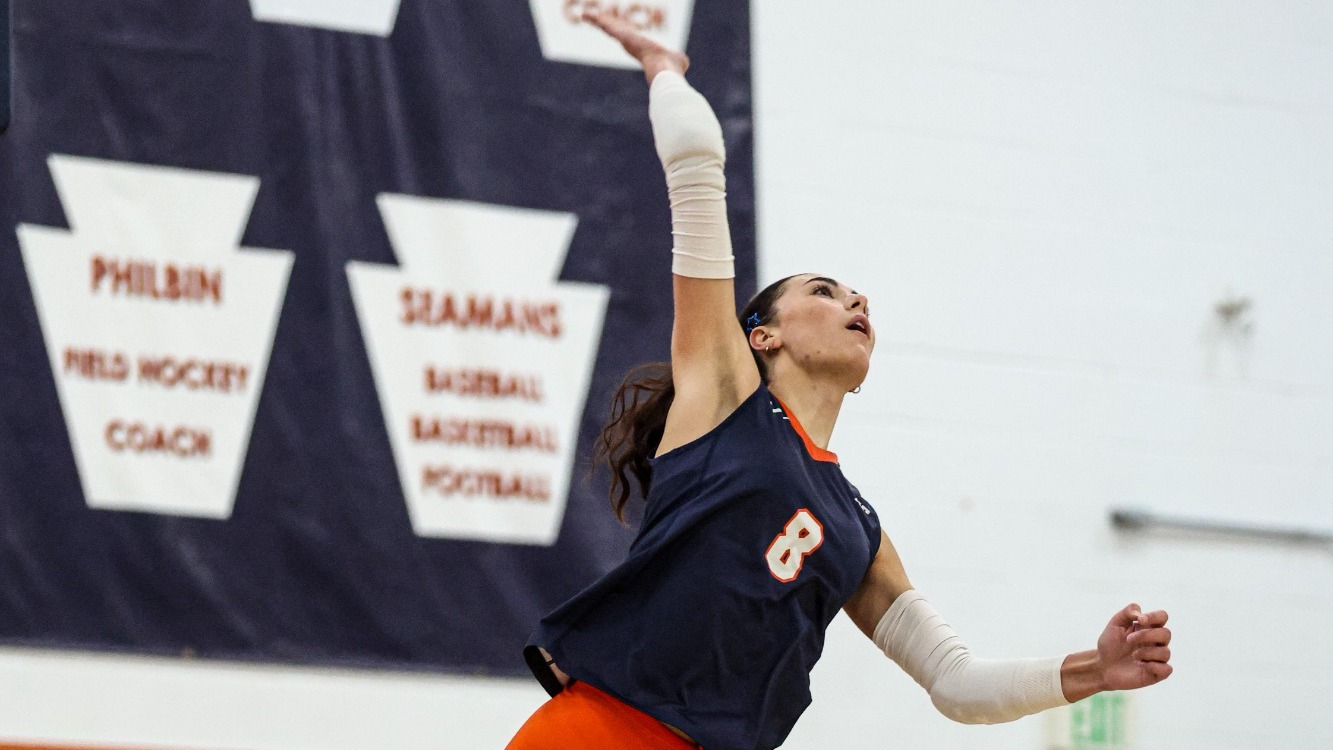 Keystone volleyball player Isabella Buonopane, wearing a navy No. 8 jersey and orange shorts, jumps to serve with her right arm extended toward the ball overhead. Teammates watch from the sideline in the background.
