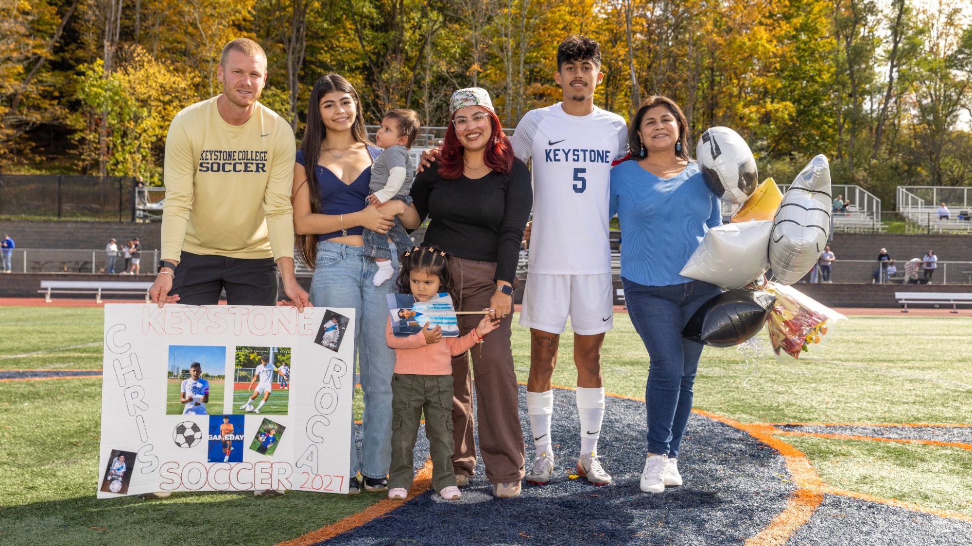 Chris Roca, wearing Keystone’s white No. 5 soccer uniform, poses with a group of adults and two children on the field during Senior Day. They display a poster with his name and soccer photos, along with soccer-themed balloons and flowers. Autumn trees fill the background.