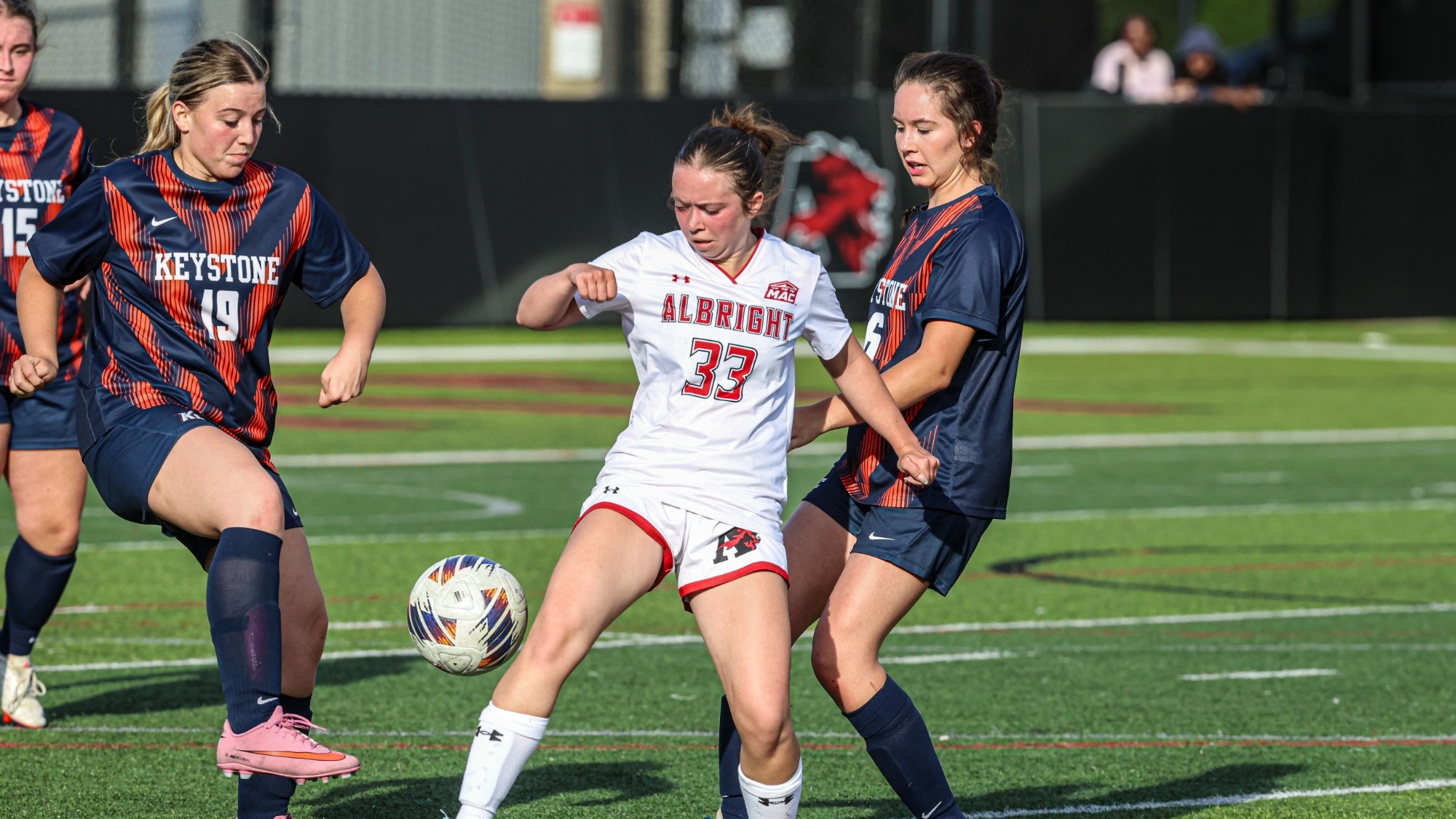 Keystone women’s soccer players in navy and orange uniforms challenge Albright’s number 33, wearing white, for an airborne ball on the turf field.