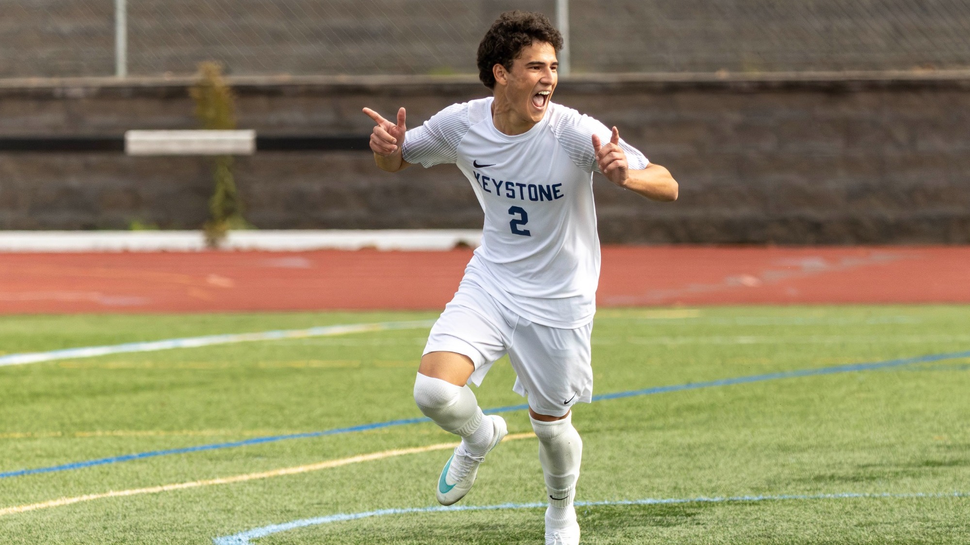 Keystone men’s soccer player wearing a white number 2 uniform runs across the turf, smiling with his mouth open and pointing both hands outward in celebration.