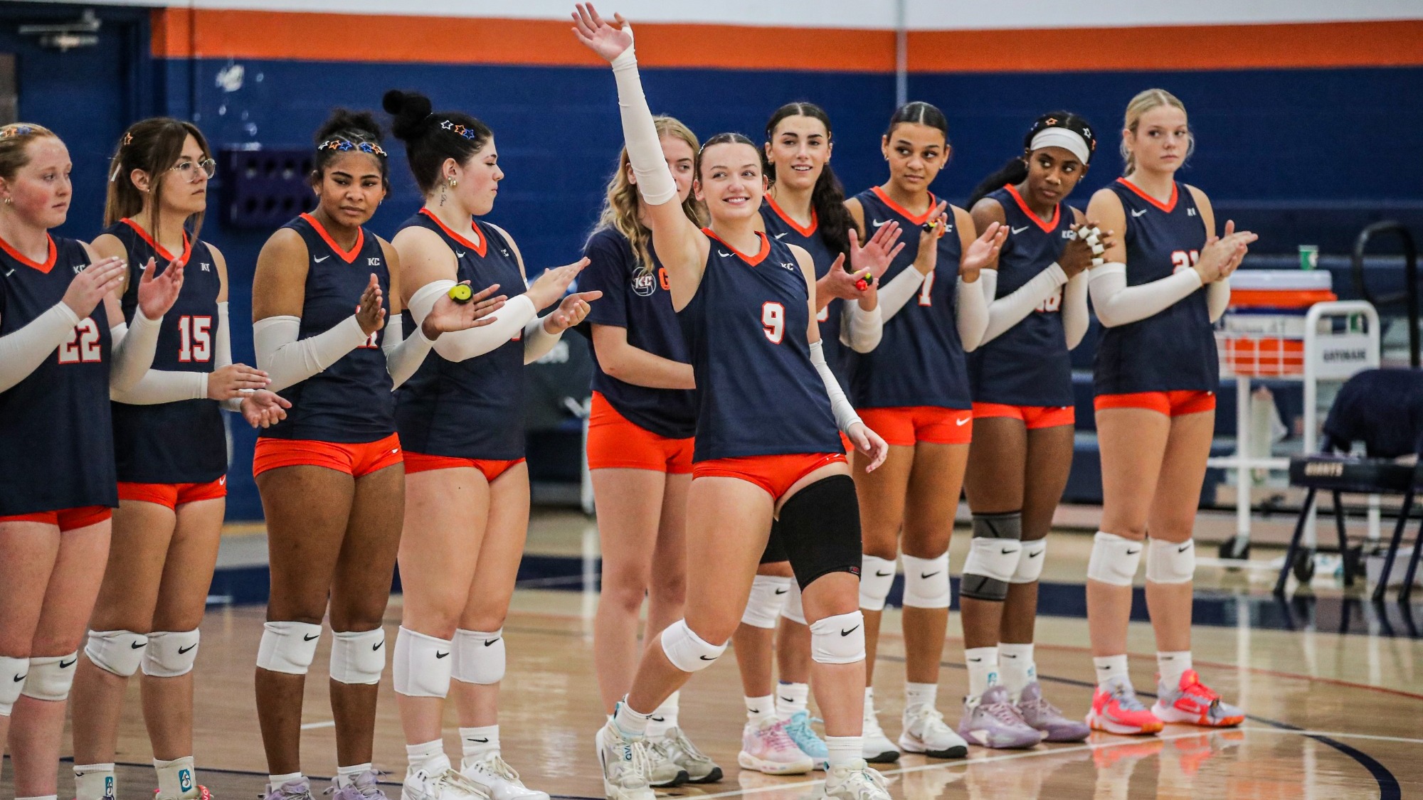  Keystone volleyball player wearing number 9 smiles and raises one hand as she steps forward from a line of applauding teammates in navy jerseys and orange shorts.