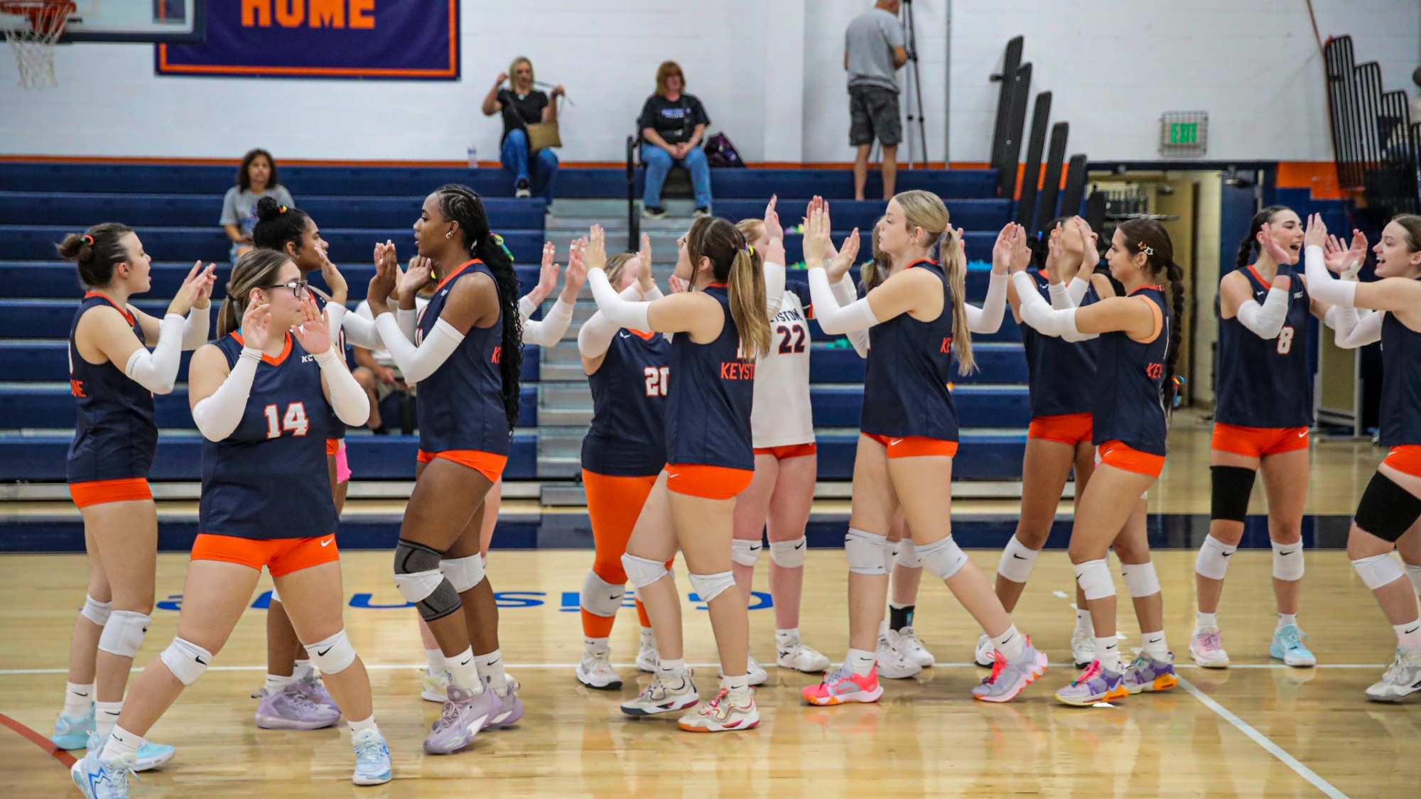 Keystone College volleyball players exchange high-fives on the court, wearing navy jerseys and orange shorts in front of the home bleachers at Gambal Athletic Center.