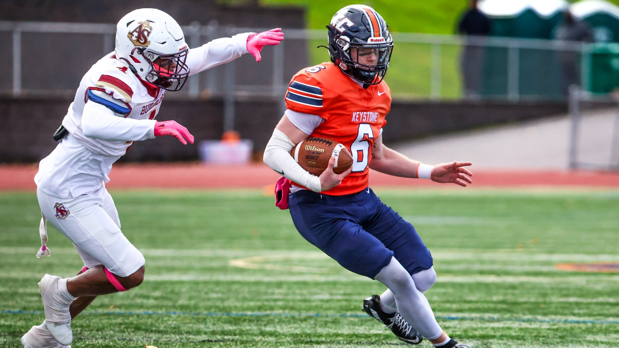 Keystone football player No. 6 carries the ball in an orange jersey and navy pants as an Apprentice defender in white pursues him across the turf.