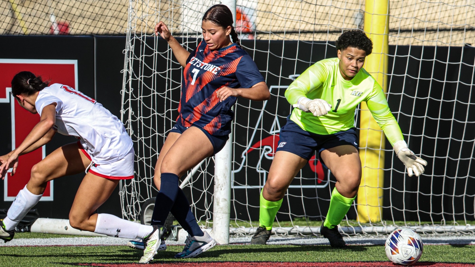 alt text please Keystone goalkeeper in a bright green jersey tracks the ball near the goal line as a teammate in navy and orange defends beside an opposing player in white.