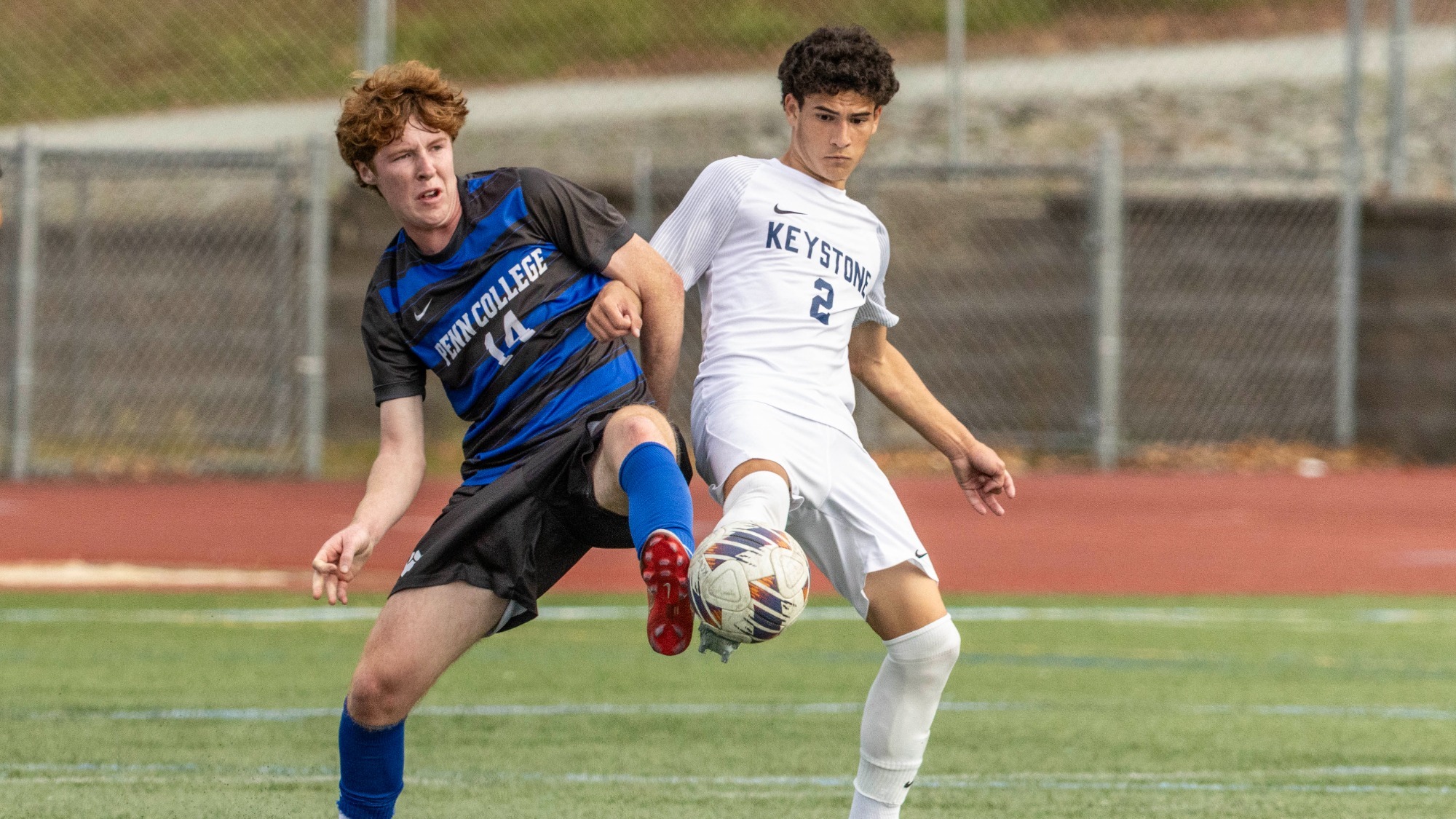  Keystone soccer player wearing No. 2 in white challenges a Penn College player in black and blue for an airborne ball on the turf field.