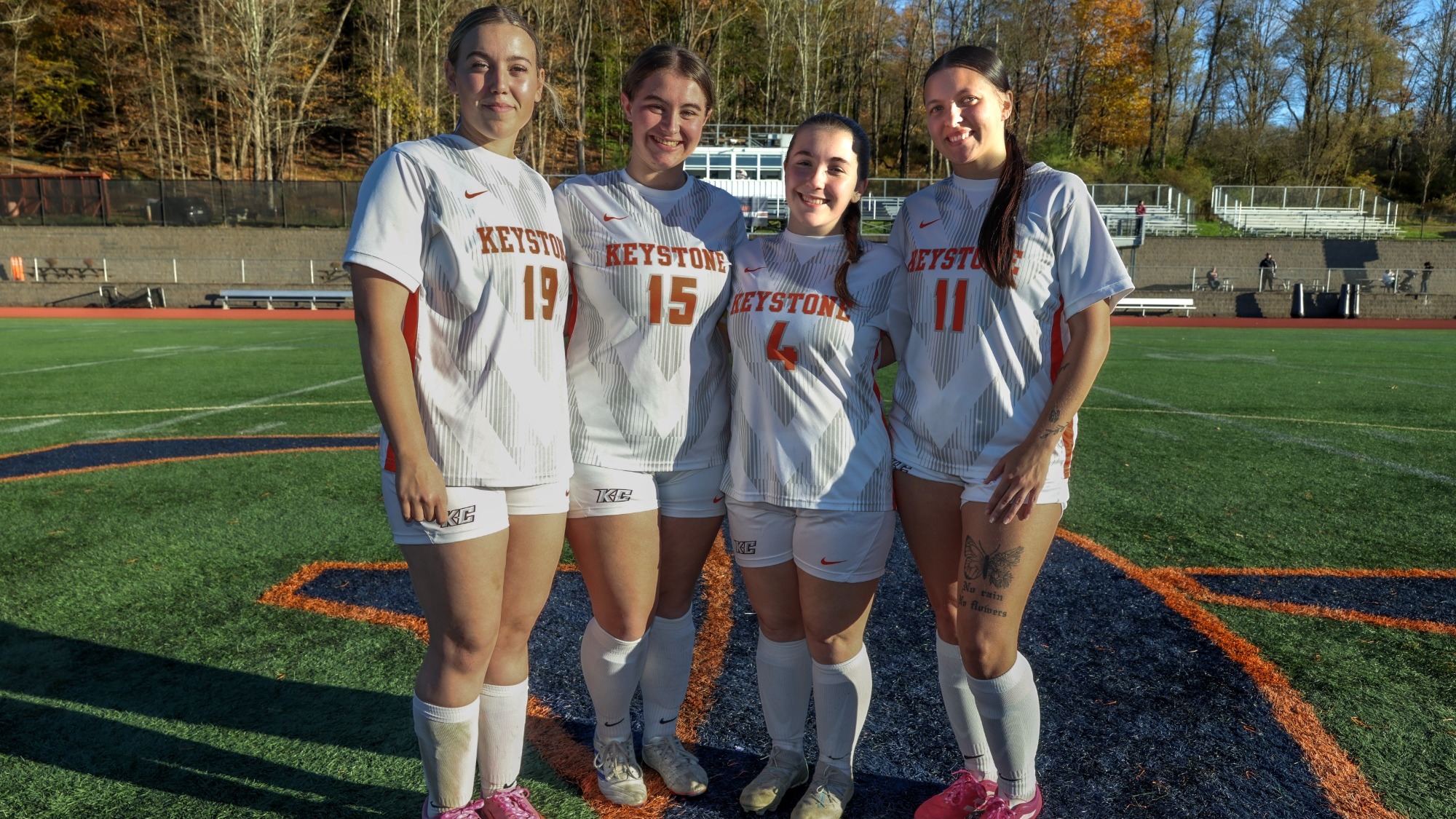 Keystone College women’s soccer seniors pose together on the field in their white Giants uniforms during Senior Day, with jersey numbers 19, 15, 4 and 11 visible. The players stand together on the midfield logo at the Keystone Turf Field and Track Complex with autumn foliage in the background.