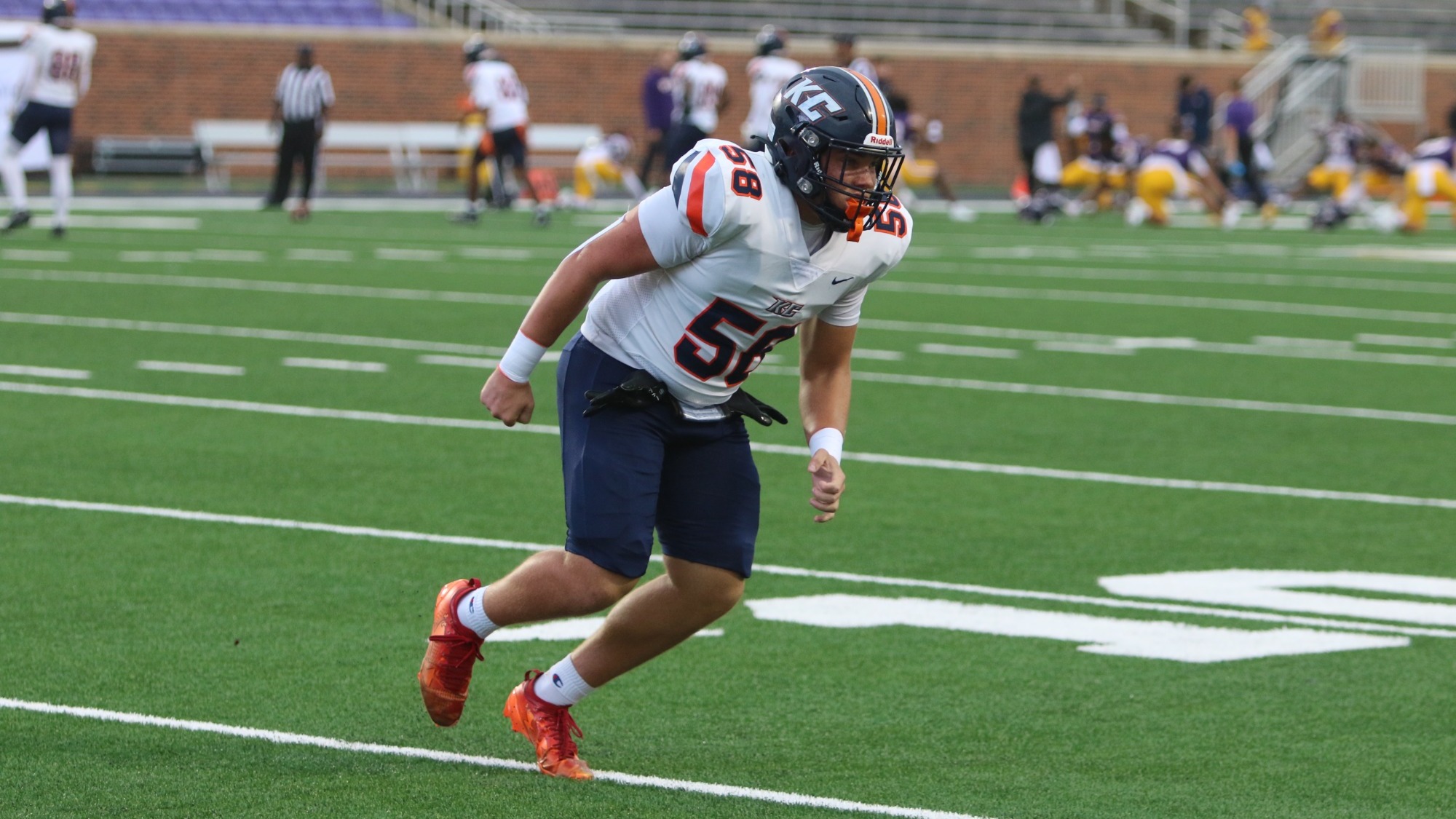  Keystone College football player No. 58 runs across the field wearing a white jersey, navy pants and helmet, and orange cleats, with players and stadium seating in the background.
