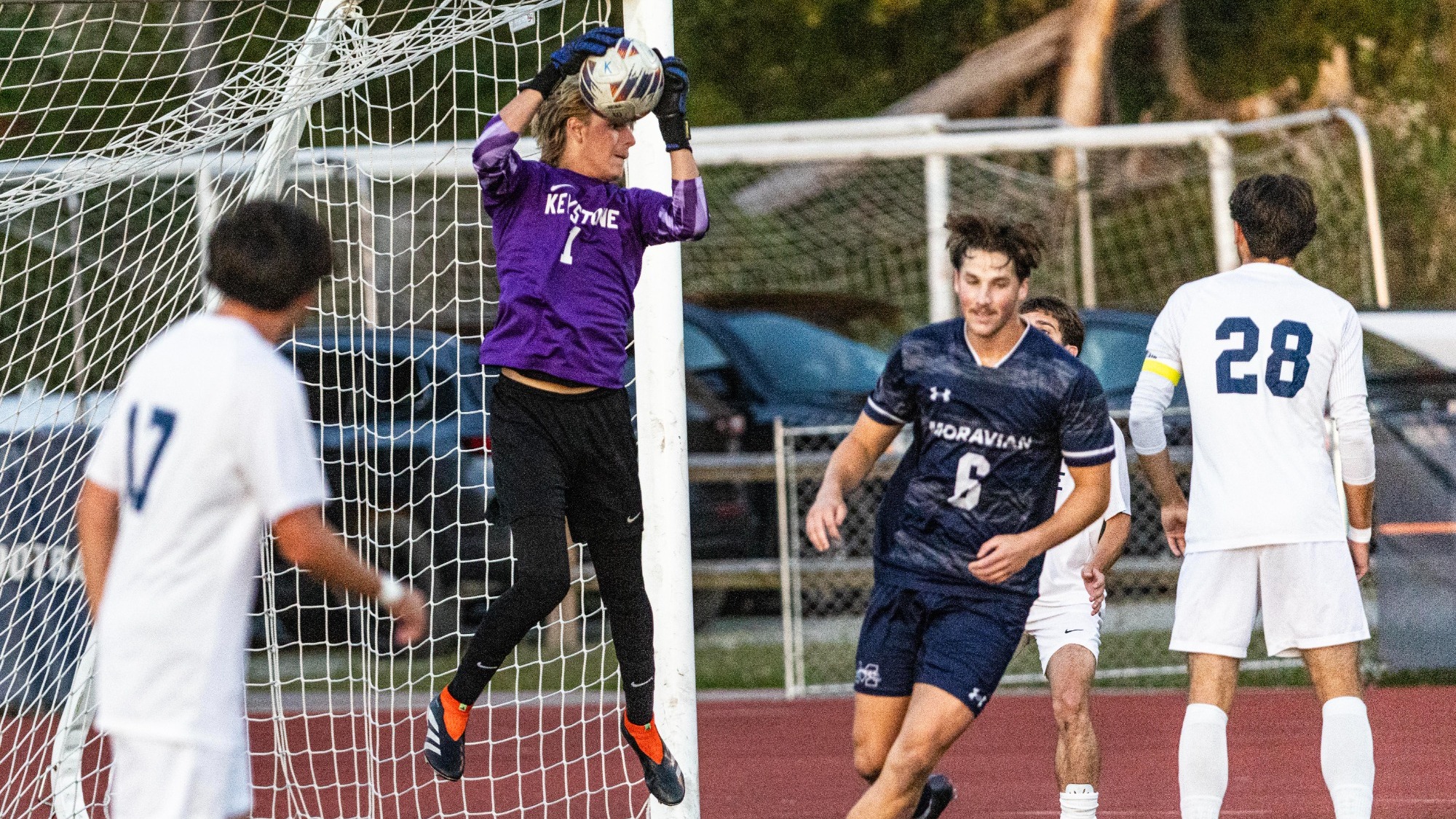 Keystone goalkeeper No. 1 jumps to catch the ball above his head near the goalpost, with teammates in white and a Moravian player nearby.